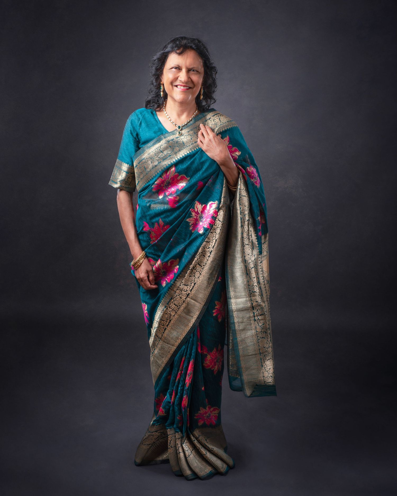 A person wears a traditional teal and gold Indian sari with floral patterns against a dark studio background.