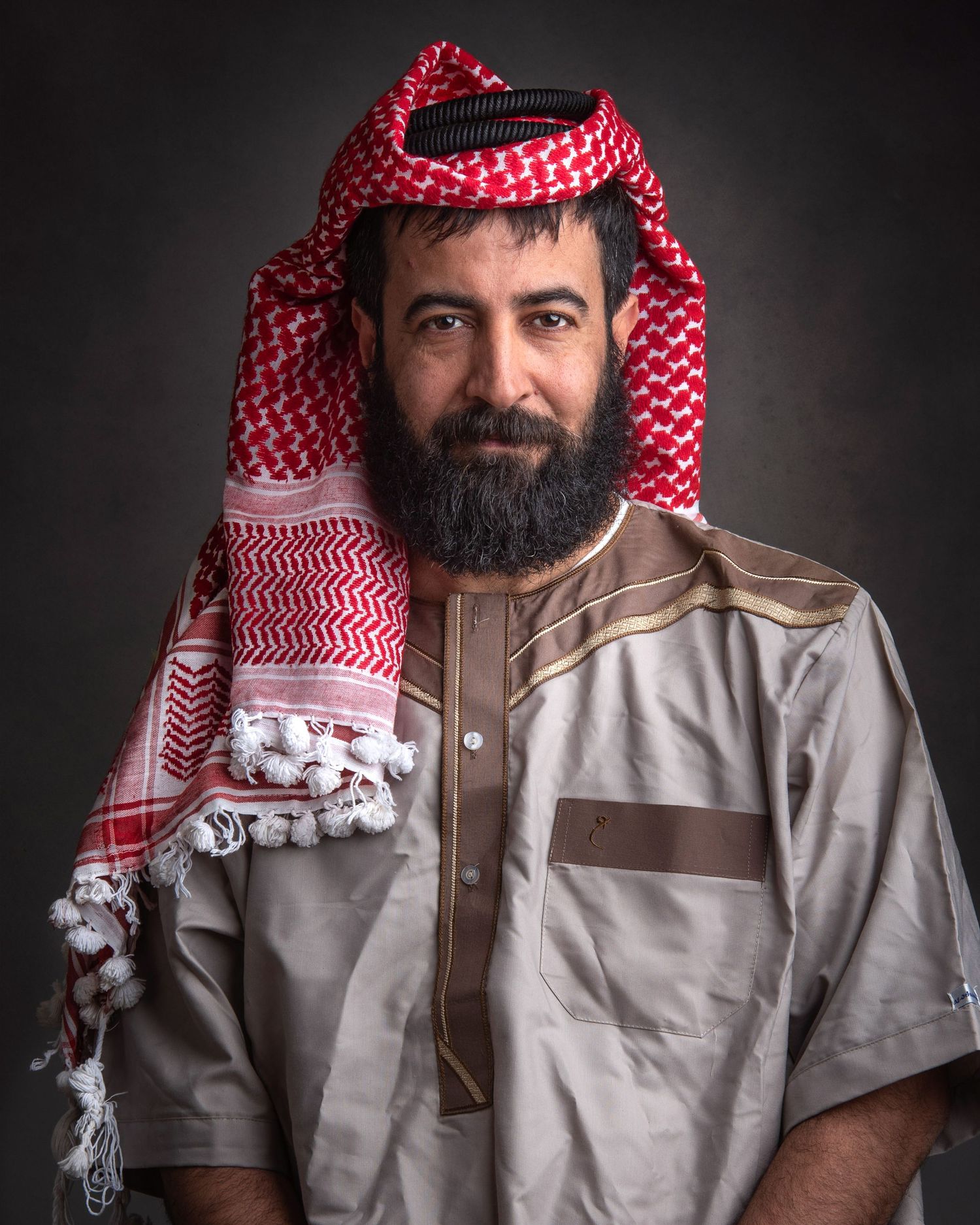 A man in traditional Middle Eastern dress including a red and white keffiyeh and light-colored thobe poses for a portrait.