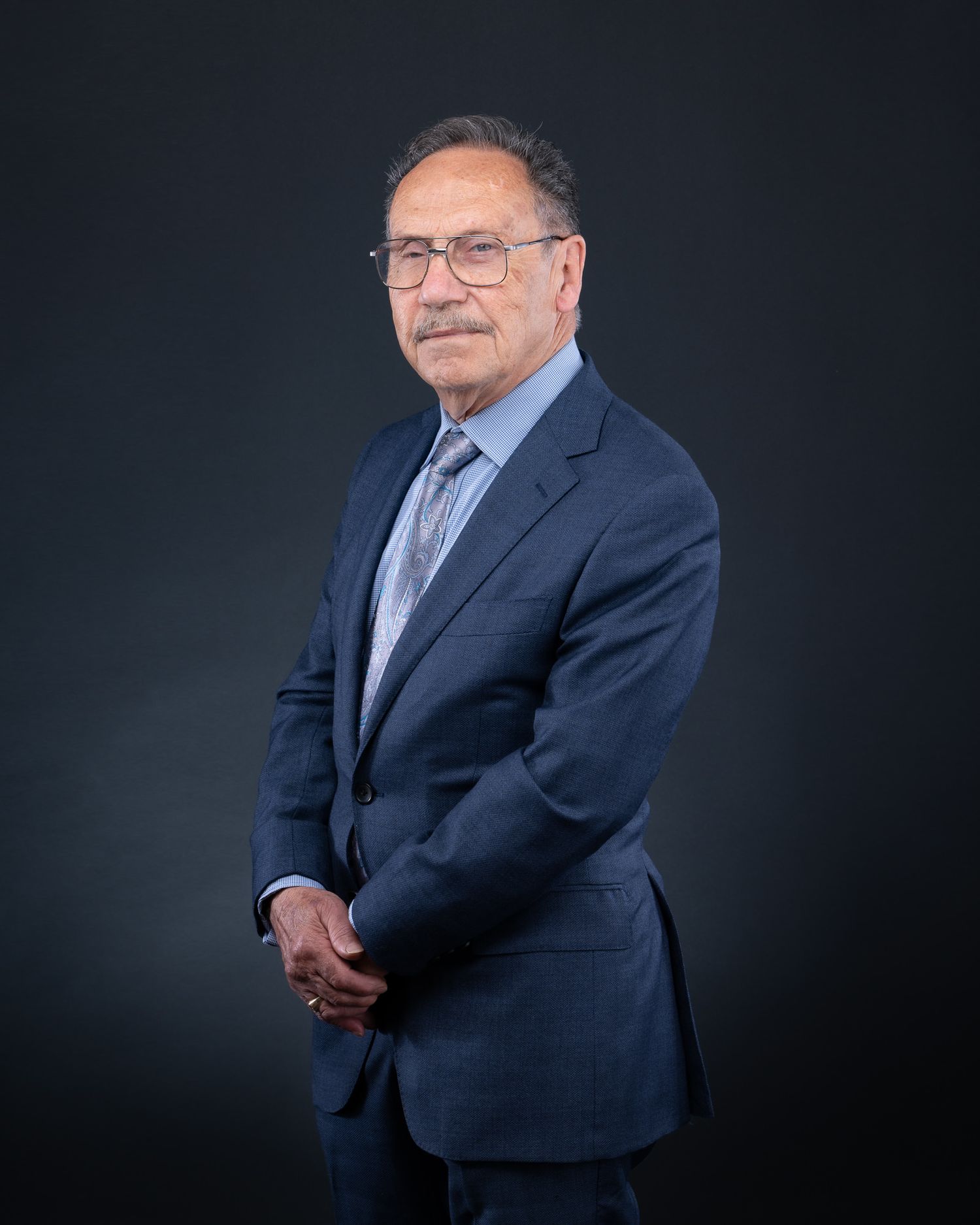 Professional studio portrait of a person in a navy suit and glasses against a dark background.