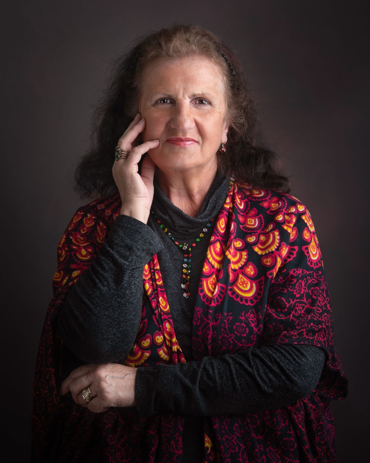 Portrait of a woman in a colorful red and black patterned sweater against a dark background with hand near face.