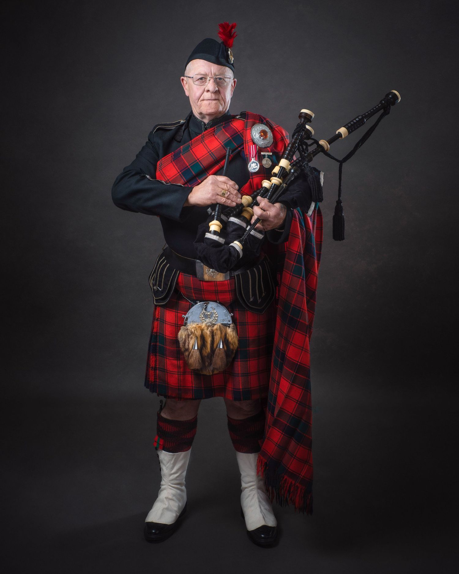 Traditional Scottish bagpiper wearing red and black tartan kilt and sash performs with bagpipes against dark background.