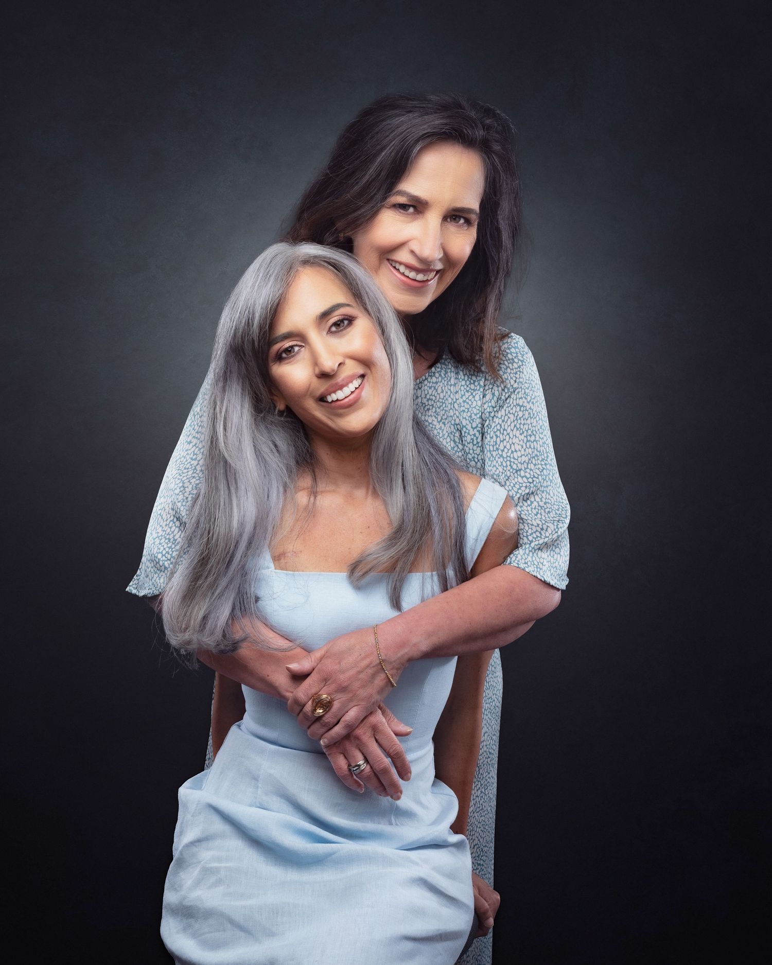 Two people embrace in an intimate portrait against a dark background wearing blue and grey tones.