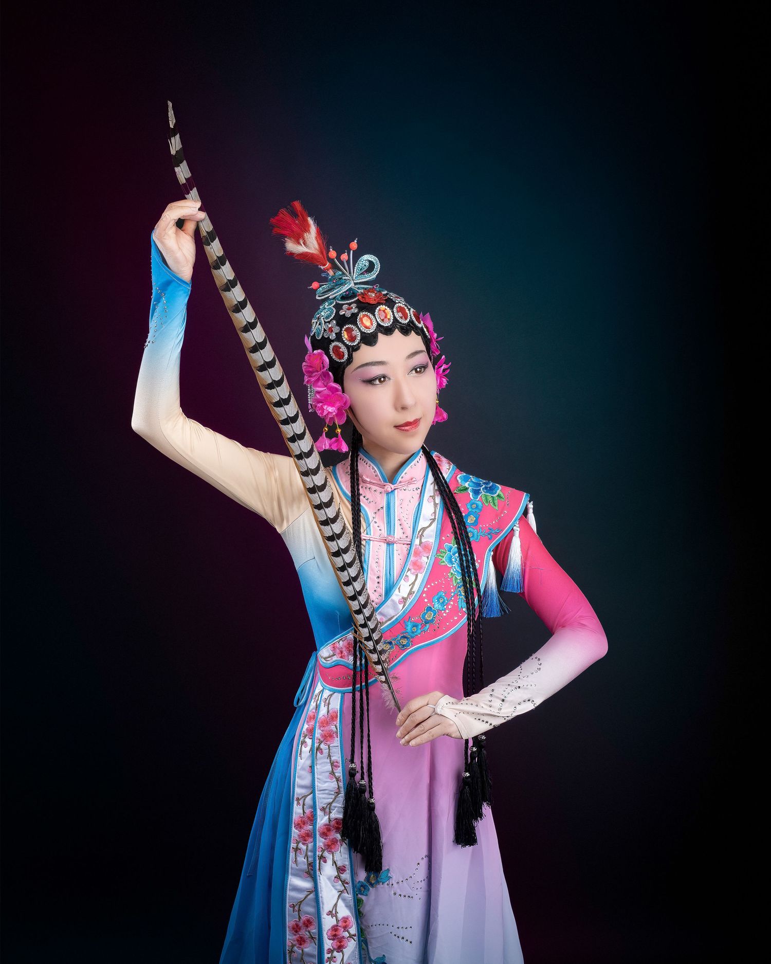 Traditional Asian performer in vibrant pink and blue hanbok dress poses dramatically against dark background with long feathers.