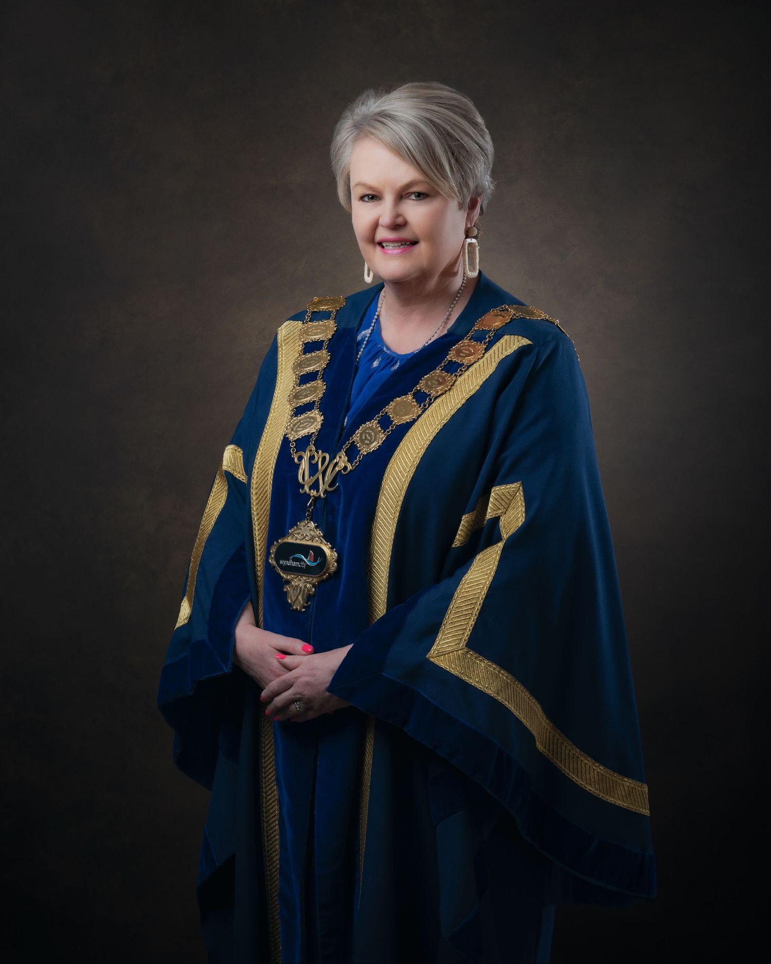 Professional studio portrait of someone wearing blue and gold ceremonial academic regalia against a dark background.