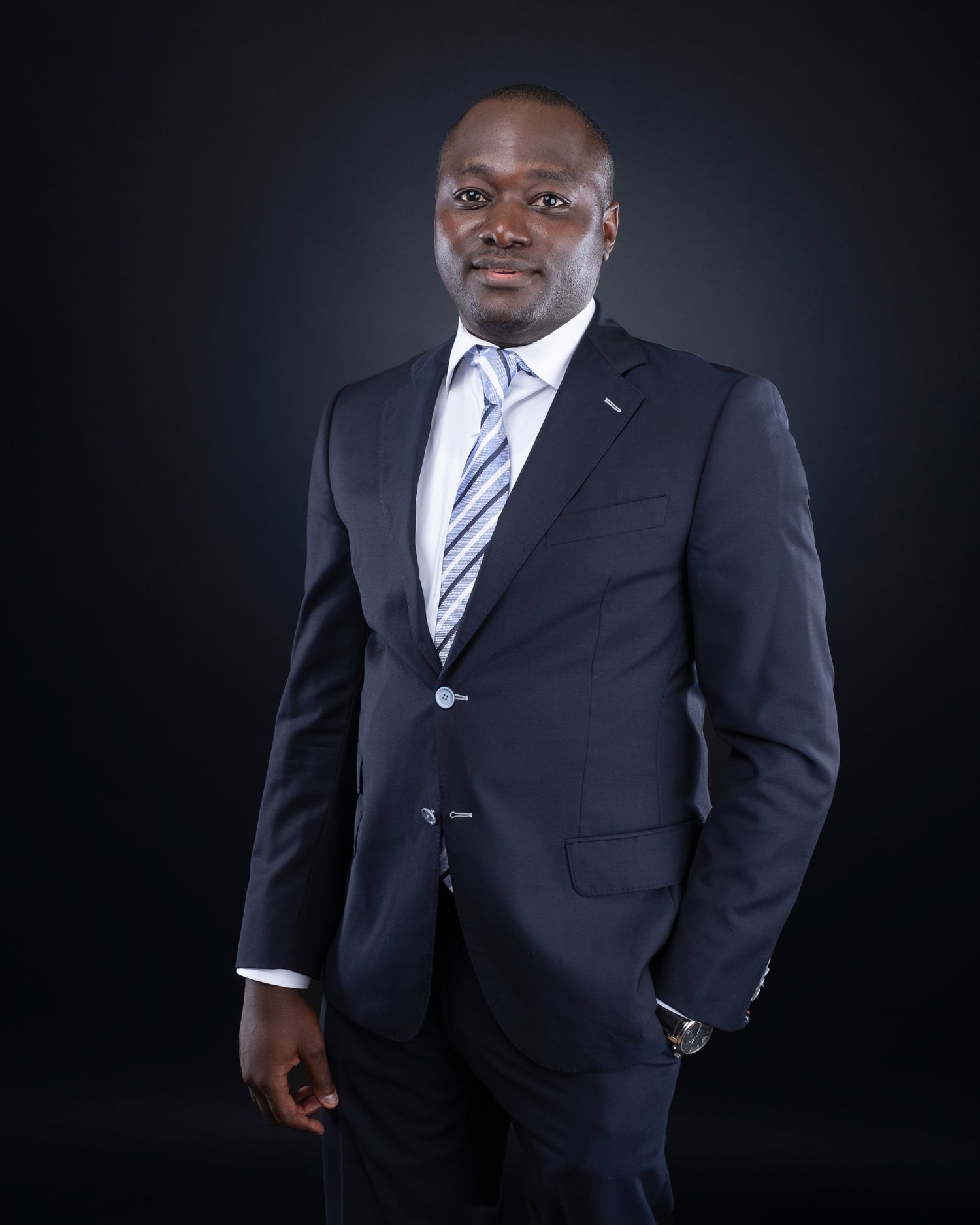 Professional headshot of person in navy suit and striped tie against dark background.