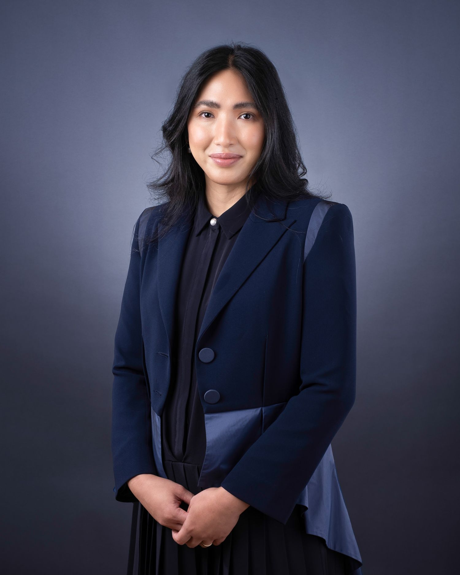 Professional studio portrait against dark background showing person in navy blazer and black top with long dark hair.