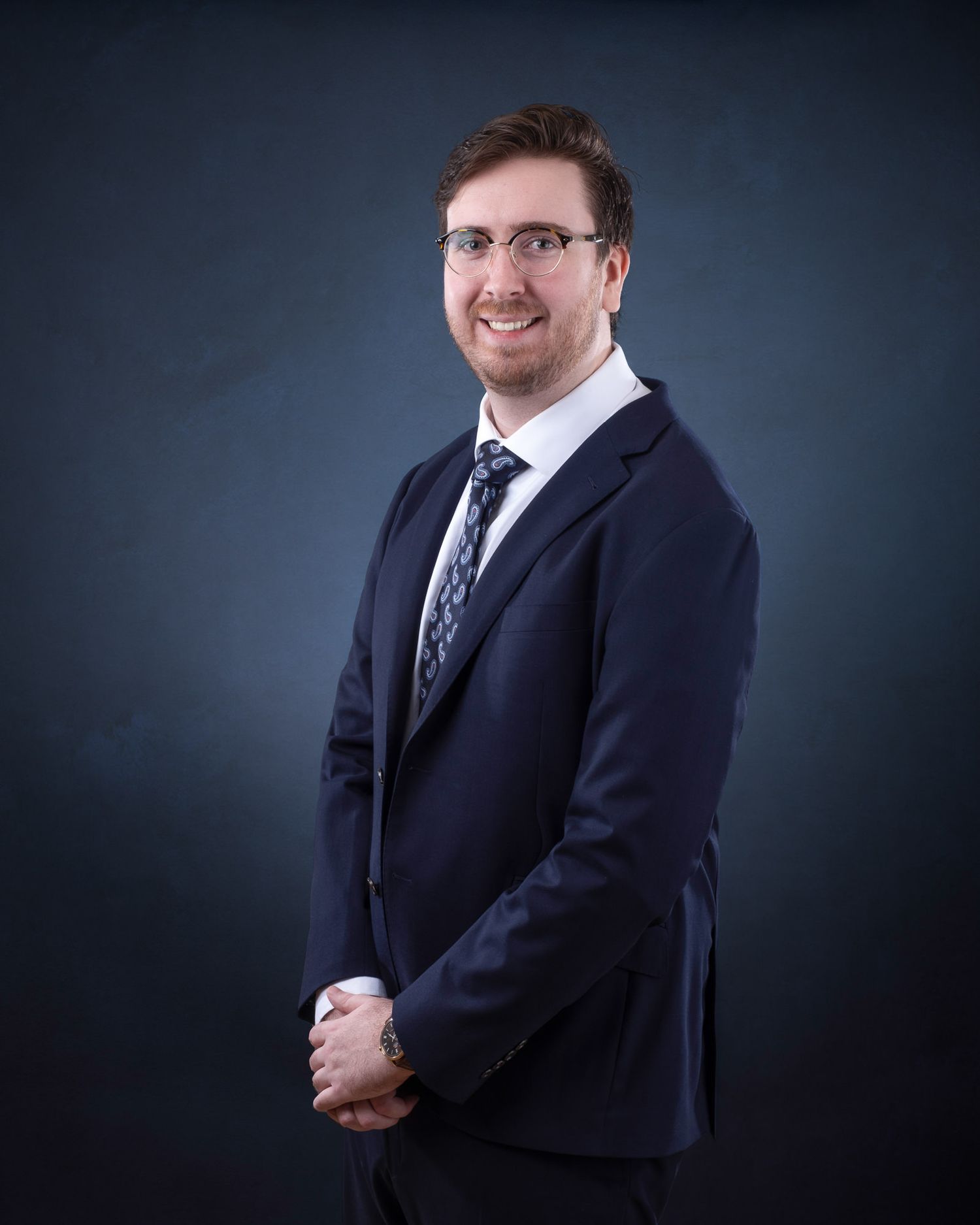 Professional headshot of someone in a navy suit and patterned tie against a dark background.