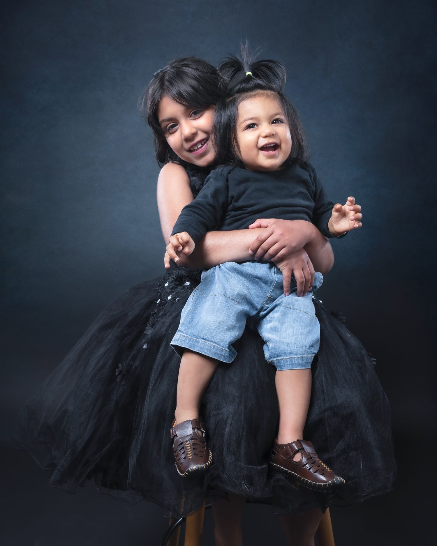 Two figures share a heartwarming embrace against a dark backdrop while wearing a black tulle dress and denim shorts with shoes.