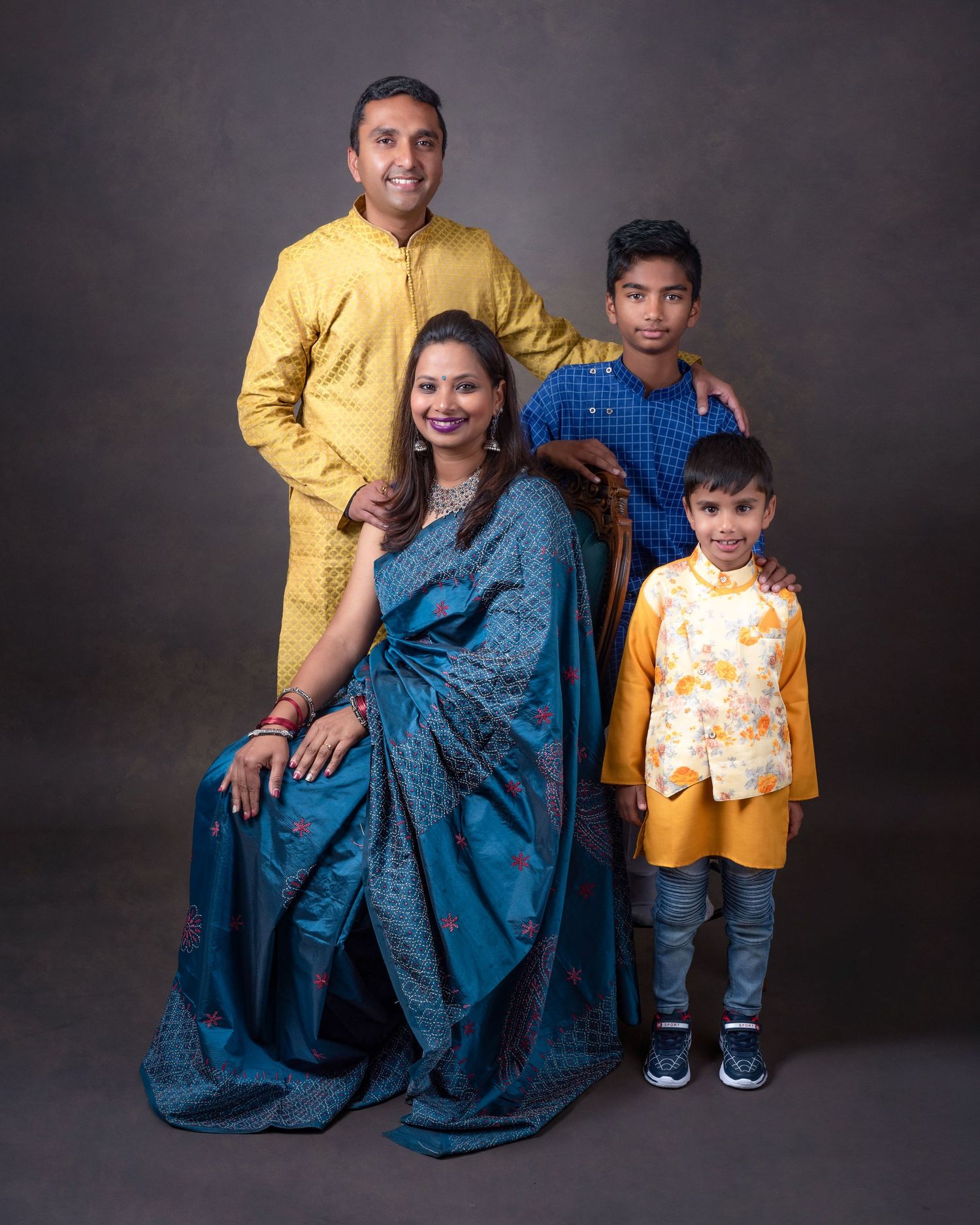 A family group dressed in traditional Indian attire poses together against a dark backdrop in a studio portrait.