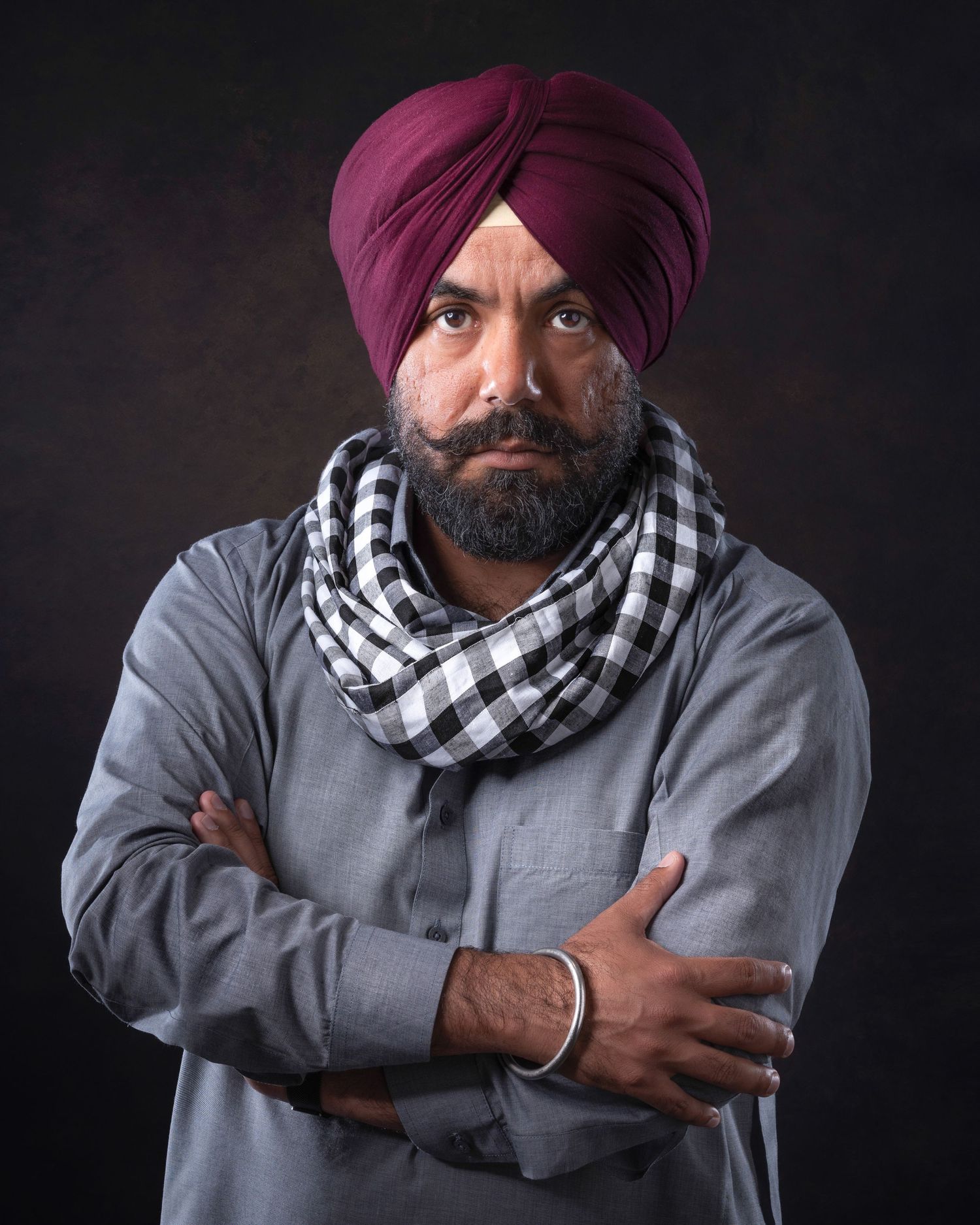 Portrait of person wearing burgundy turban and checkered scarf with gray jacket against dark background.