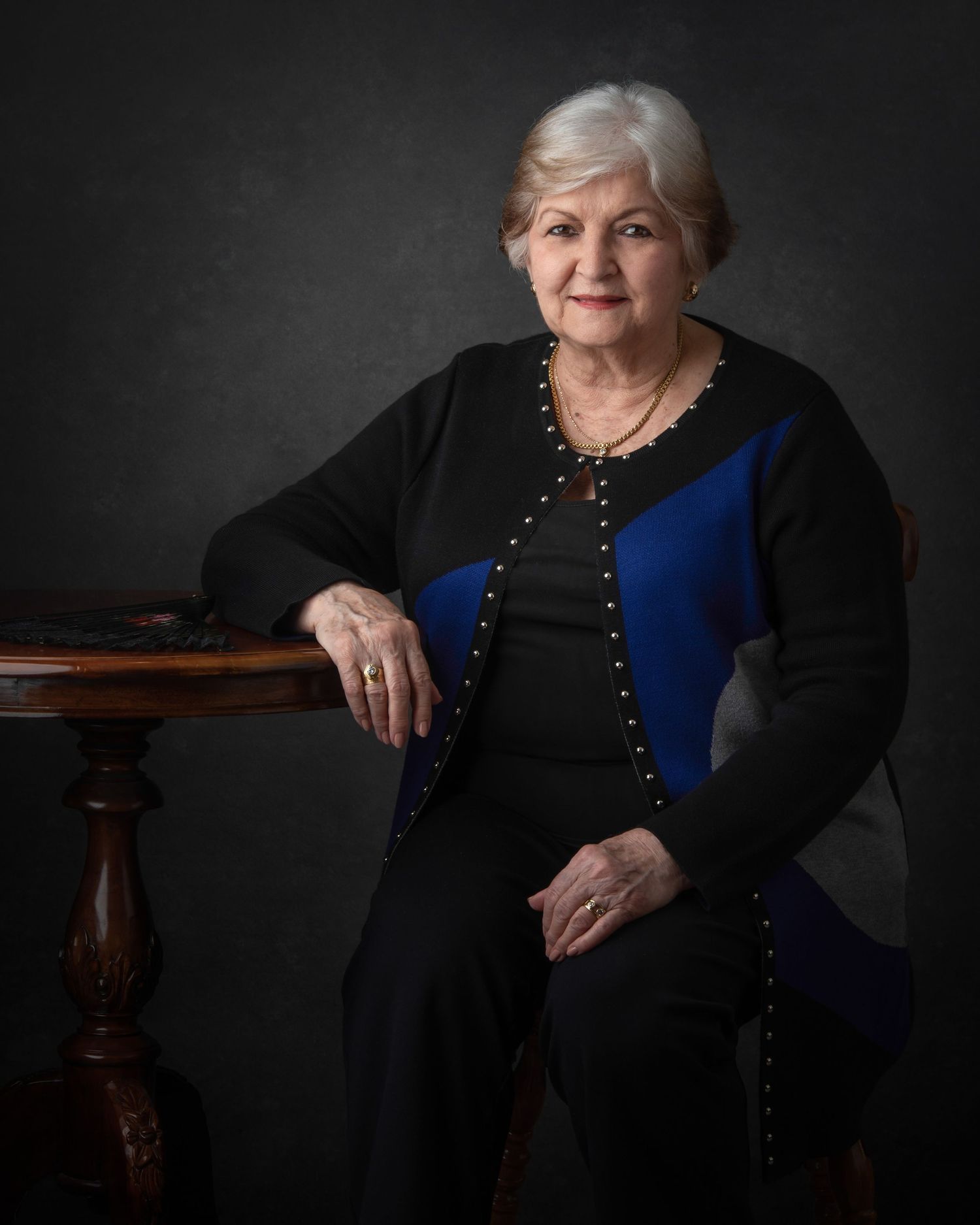 Elegant formal portrait against dark backdrop featuring black and royal blue dress with ornate detailing and antique wooden table.