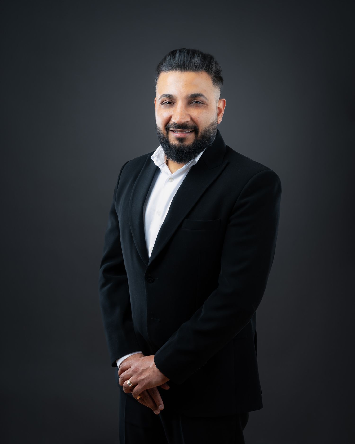 Professional business headshot of a person wearing a black blazer and white shirt against a dark backdrop.