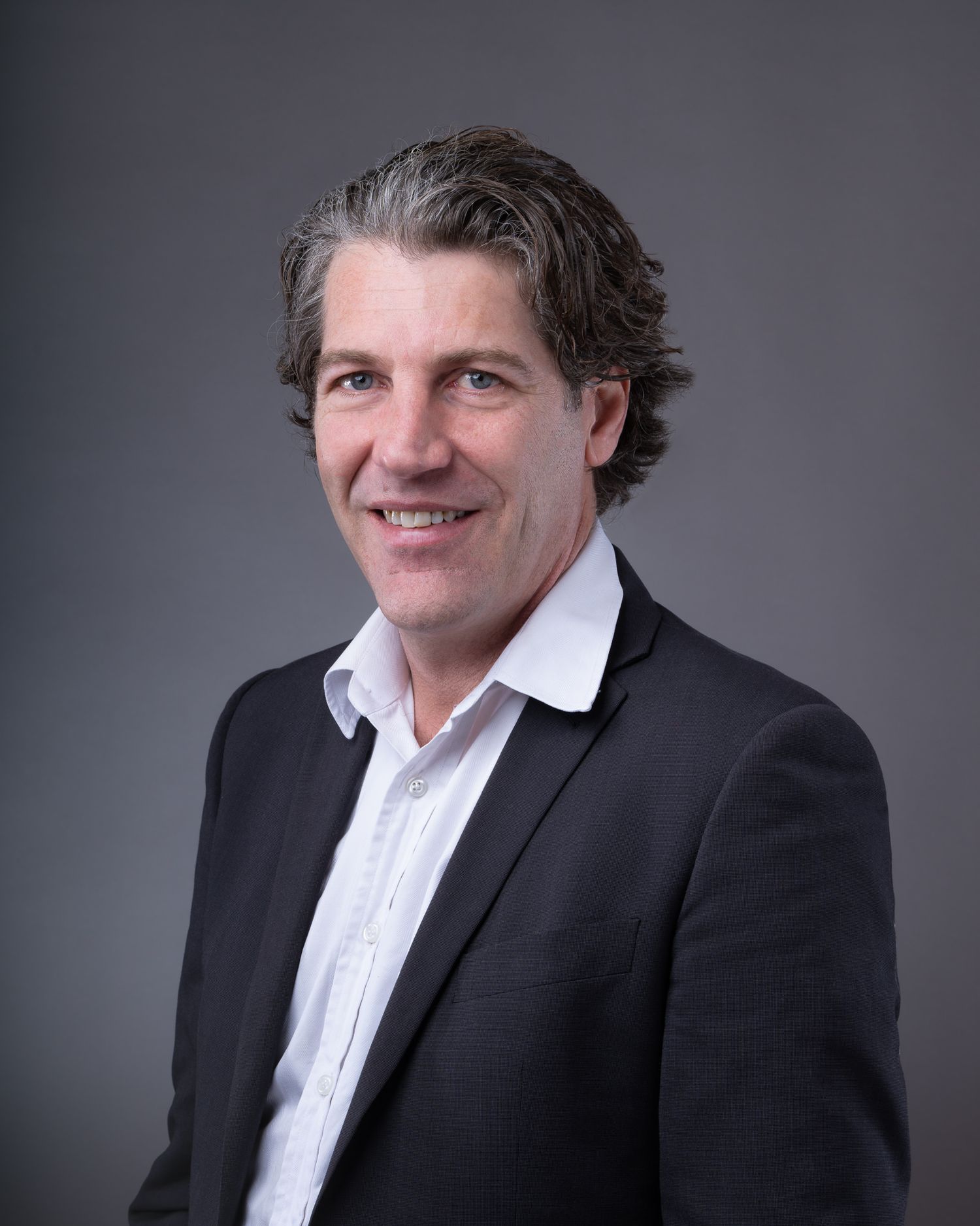 Professional business portrait of person in black suit jacket and white collared shirt against gray background.
