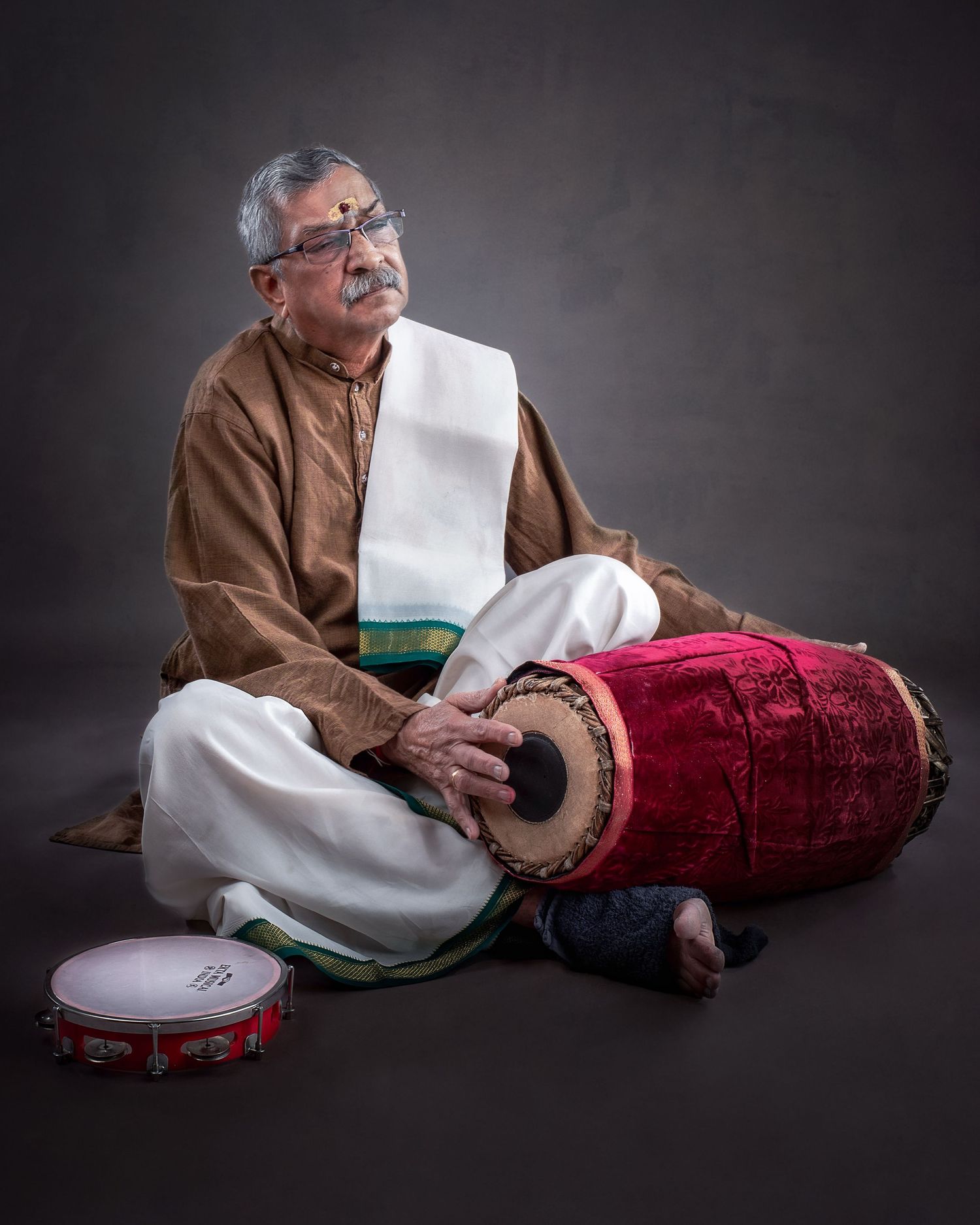 A drummer in traditional white lungi and brown kurta plays mridangam and tambourine against a dark studio backdrop.