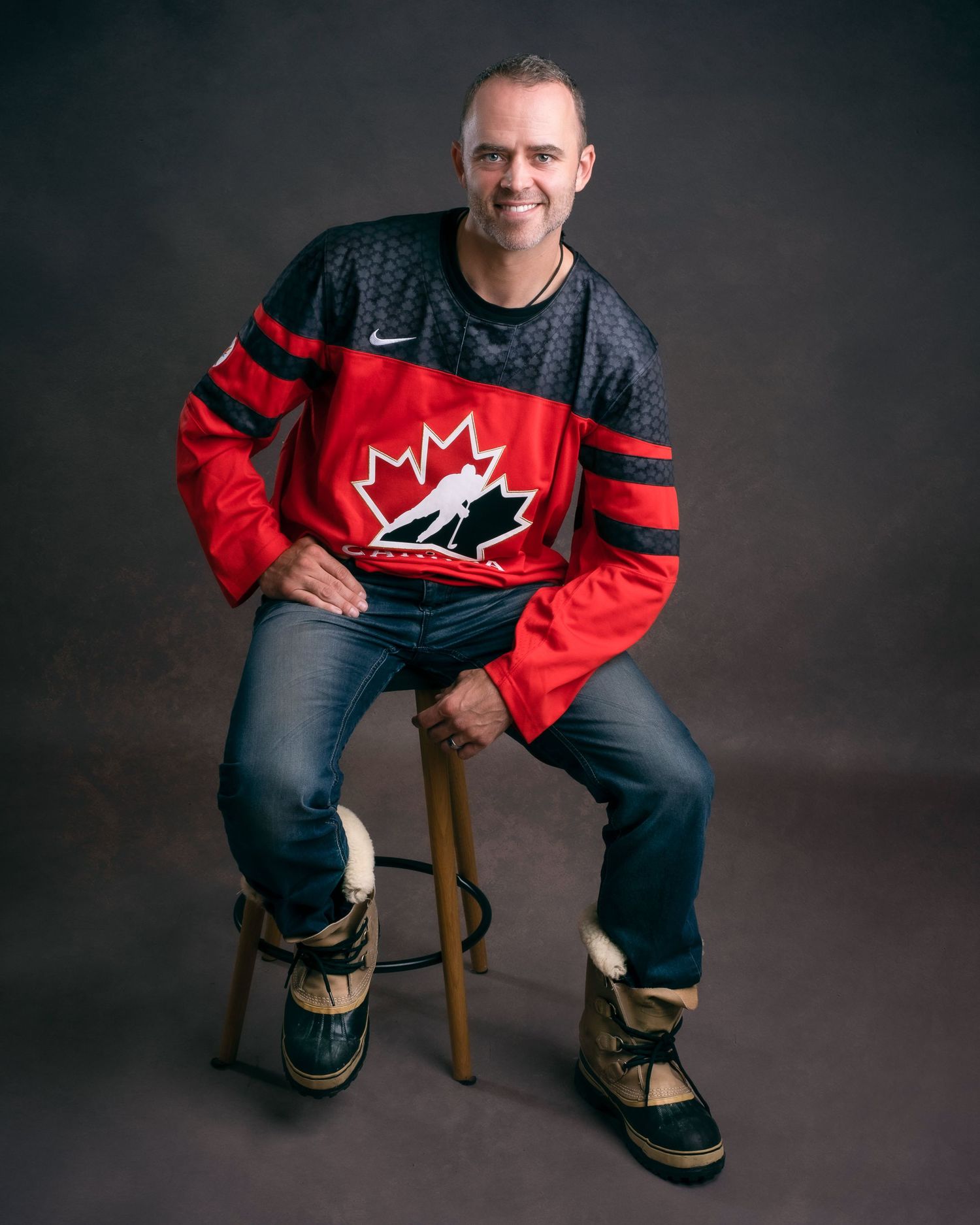 Someone in blue jeans and a Team Canada hockey jersey sits on a stool against a dark studio backdrop.