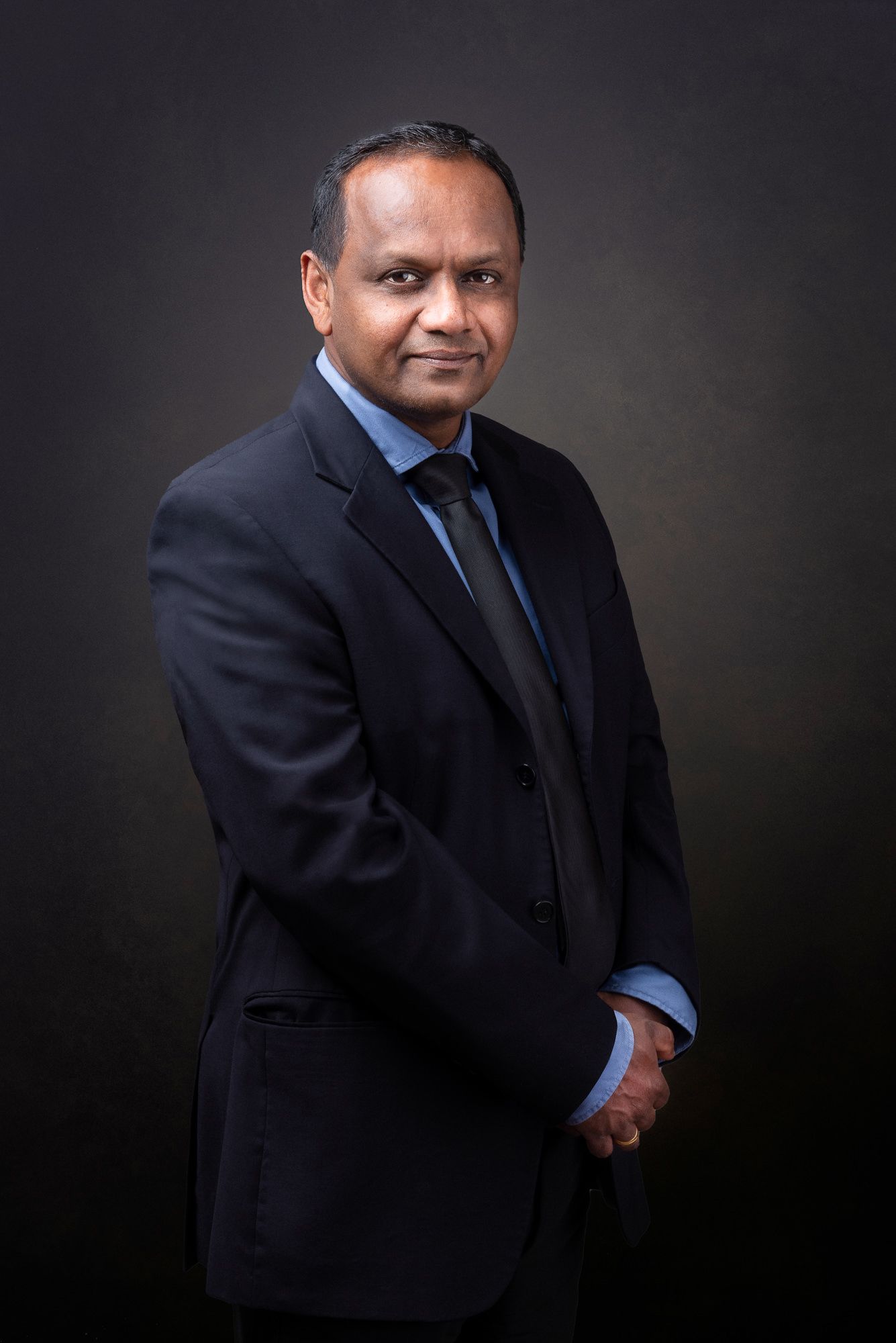Professional studio portrait of someone in a navy suit and blue tie against a dark background with dramatic lighting.