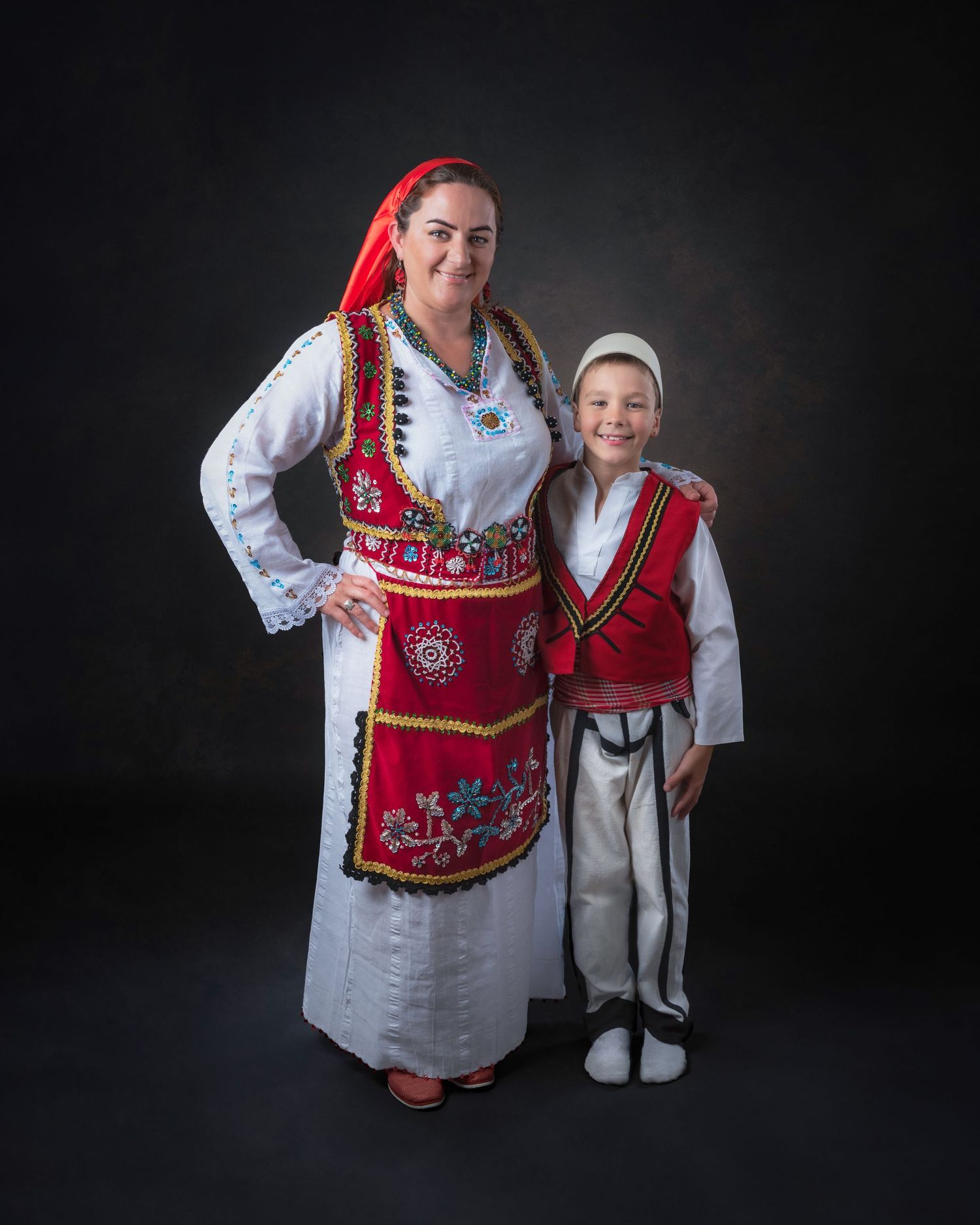 Two people wearing traditional Norwegian folk costumes with red and white colors pose together against dark backdrop.