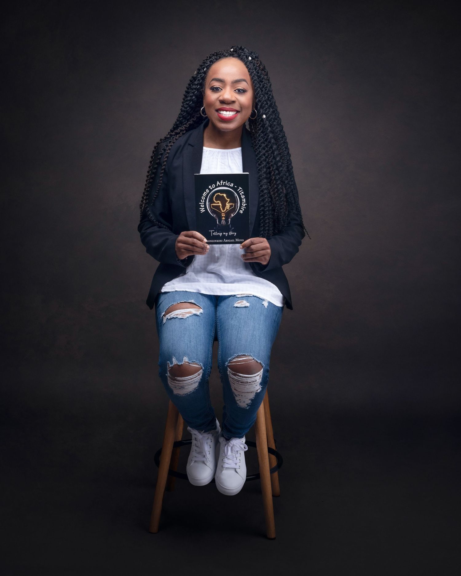 A smiling person wearing braids sits on a wooden stool against a dark background, wearing ripped jeans and holding a book.