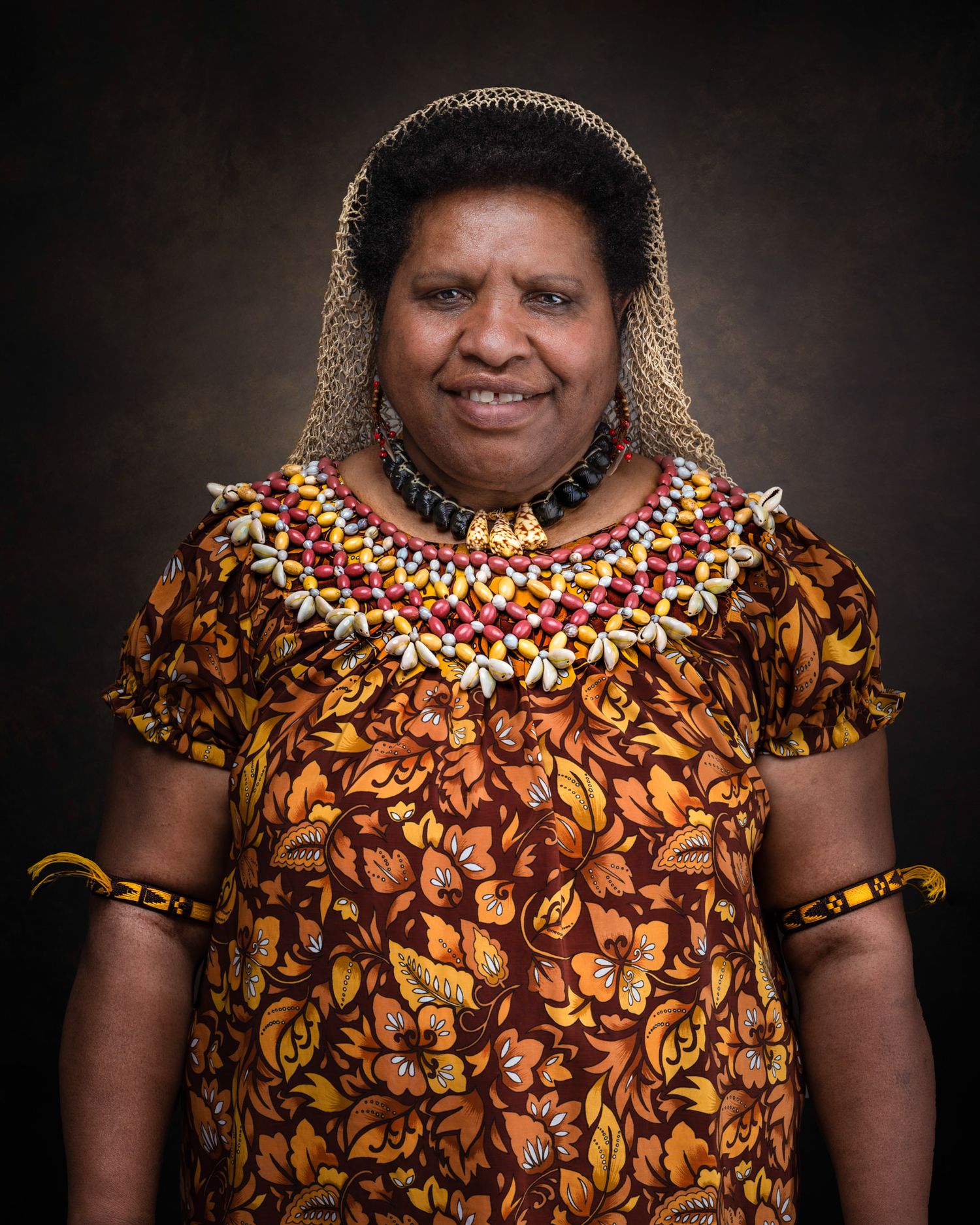 Portrait of a person wearing vibrant traditional Pacific Islander clothing with intricate beaded necklace against dark background.