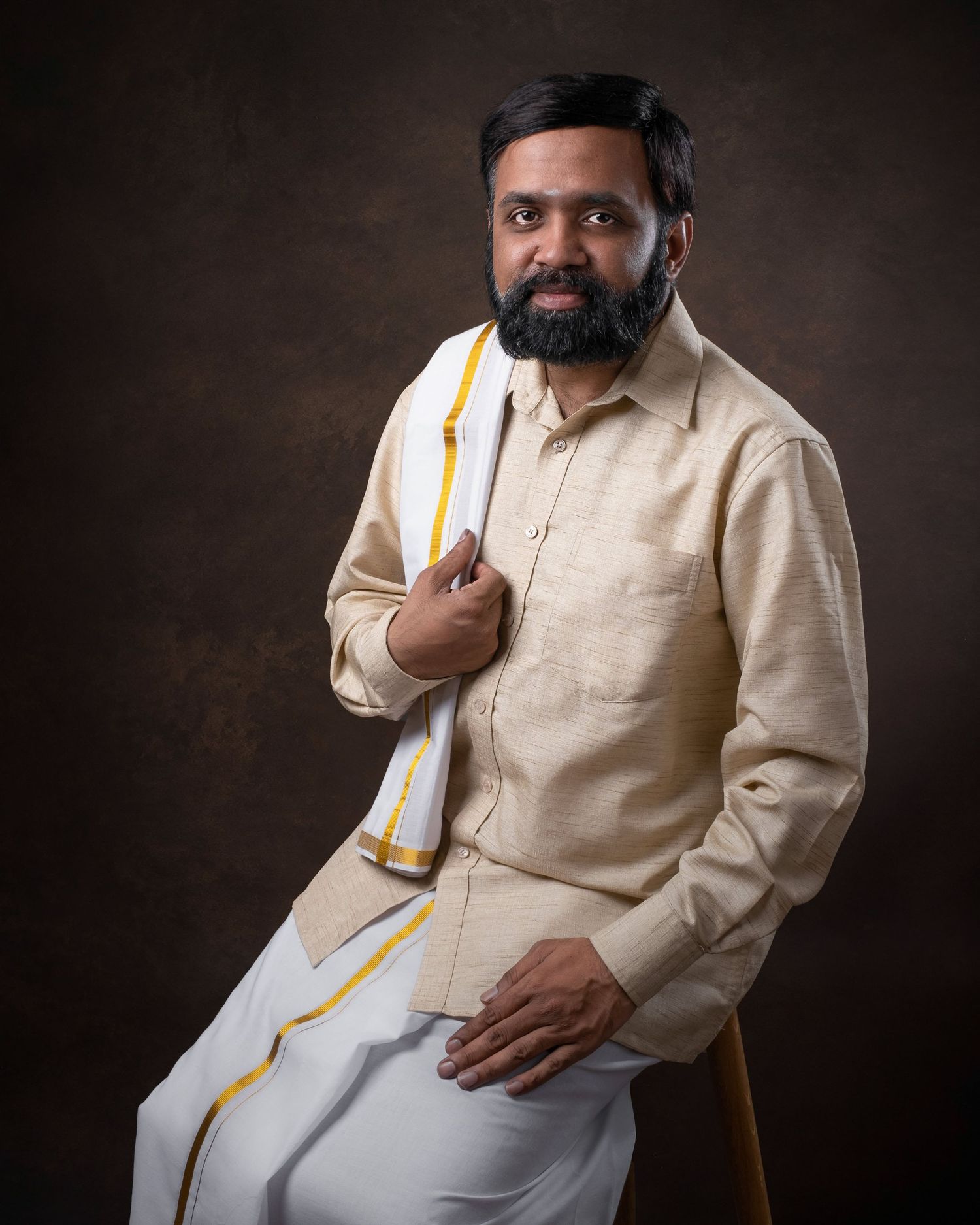 Professional portrait of someone in traditional Indian attire with a cream shirt and white dhoti against dark background.