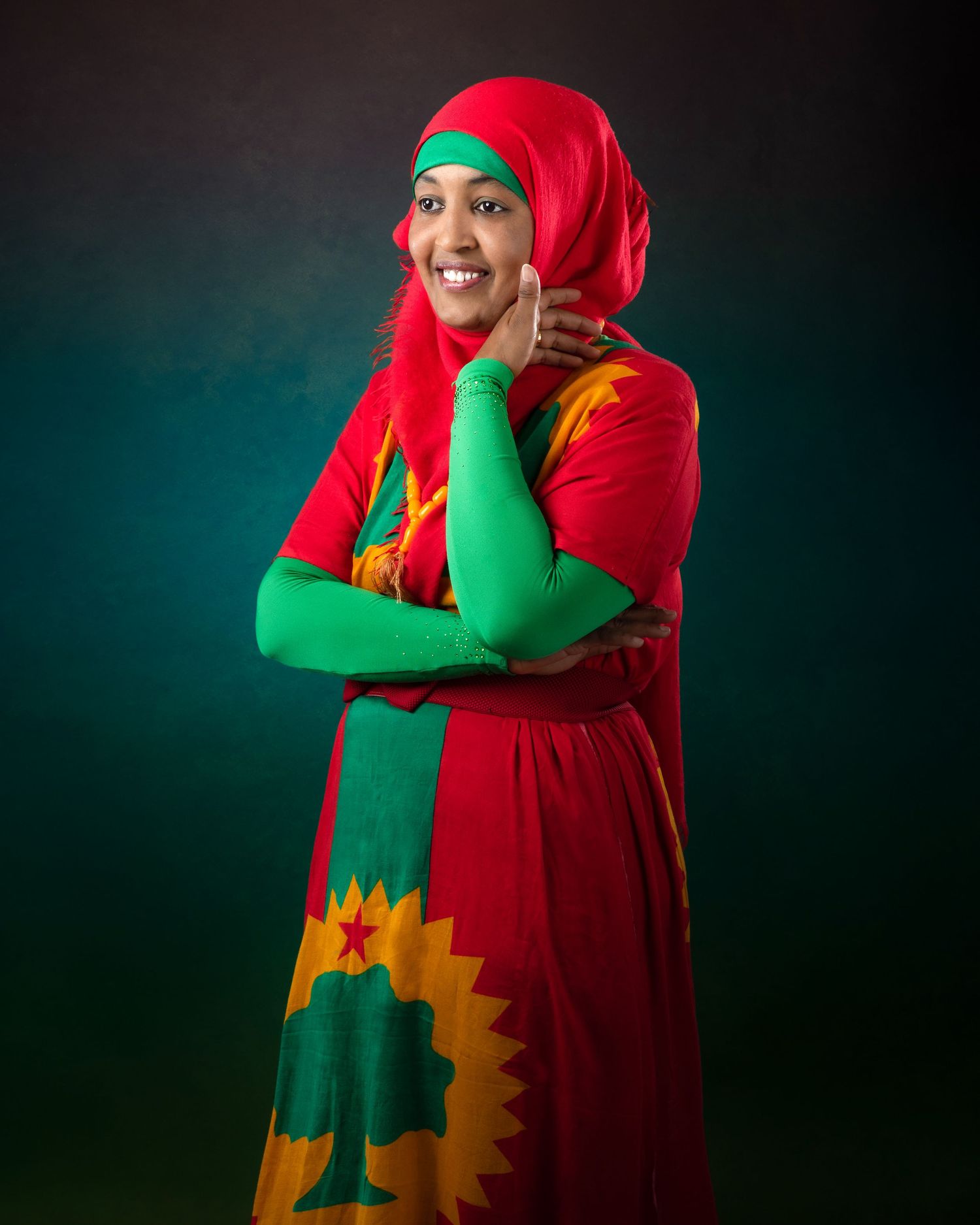 A person wearing vibrant red and green traditional attire with decorative patterns poses joyfully against a dark background.