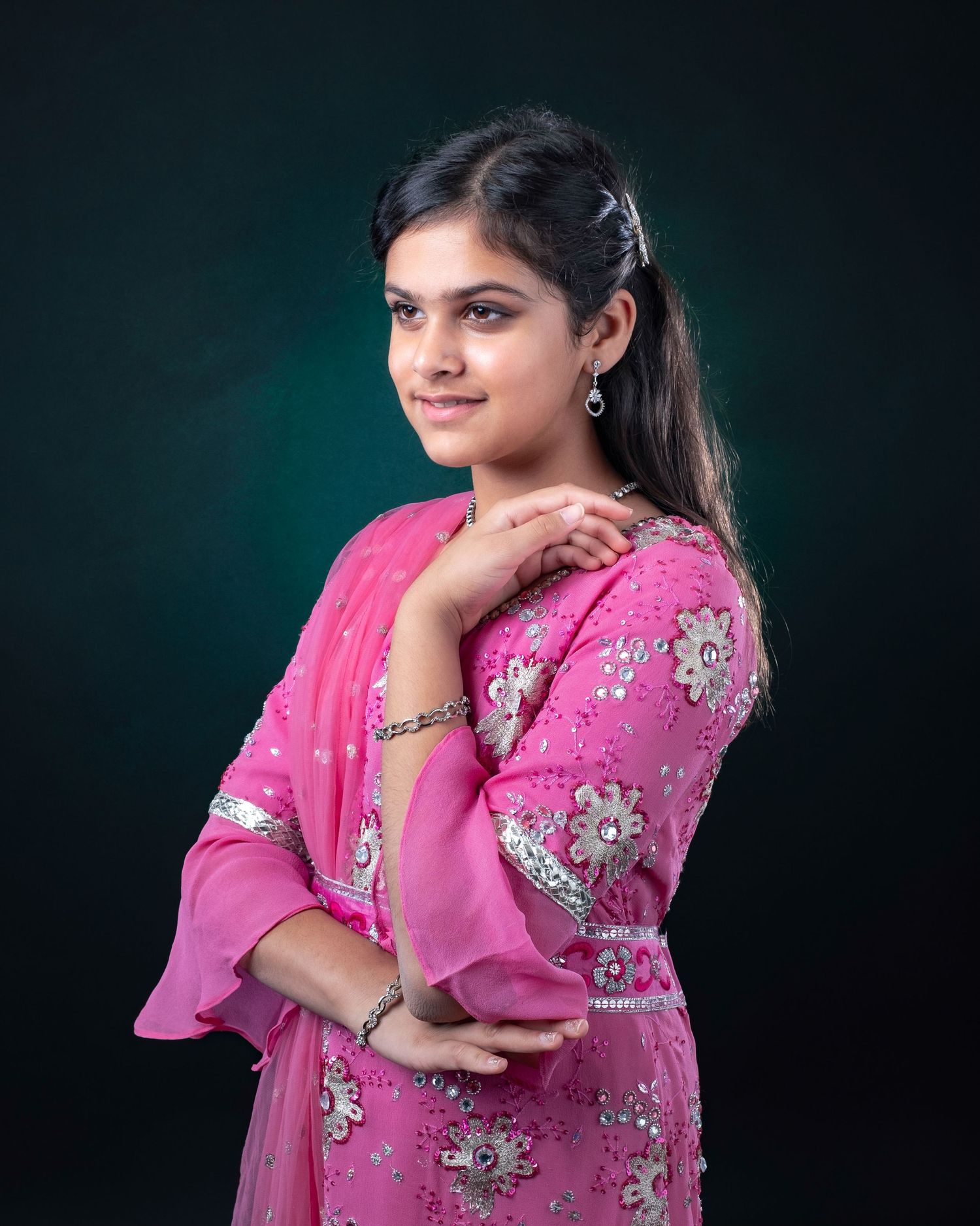 A person wearing a pink floral embellished dress poses against a dark green studio backdrop with hand near shoulder.