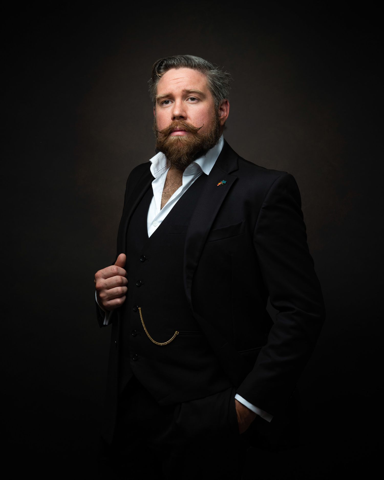Professional portrait against dark background of person in three-piece suit with beard wearing watch chain and white shirt.