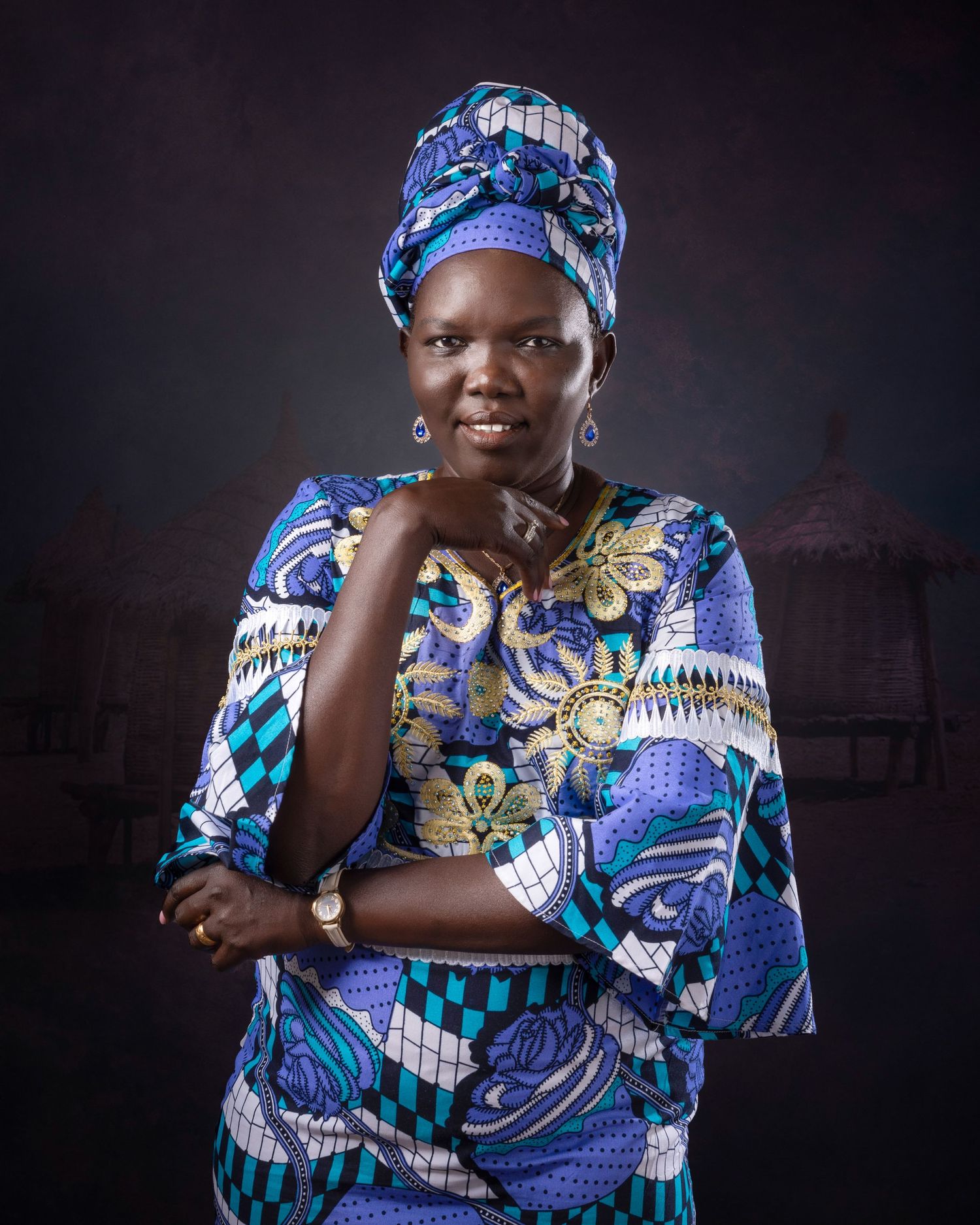 Person wearing vibrant blue and white traditional African print dress with matching head wrap poses against dark background.