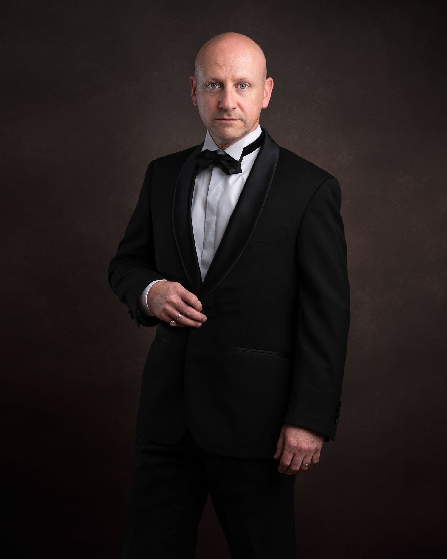A formal portrait of someone wearing a black tuxedo with bow tie against a dark background in classic style.