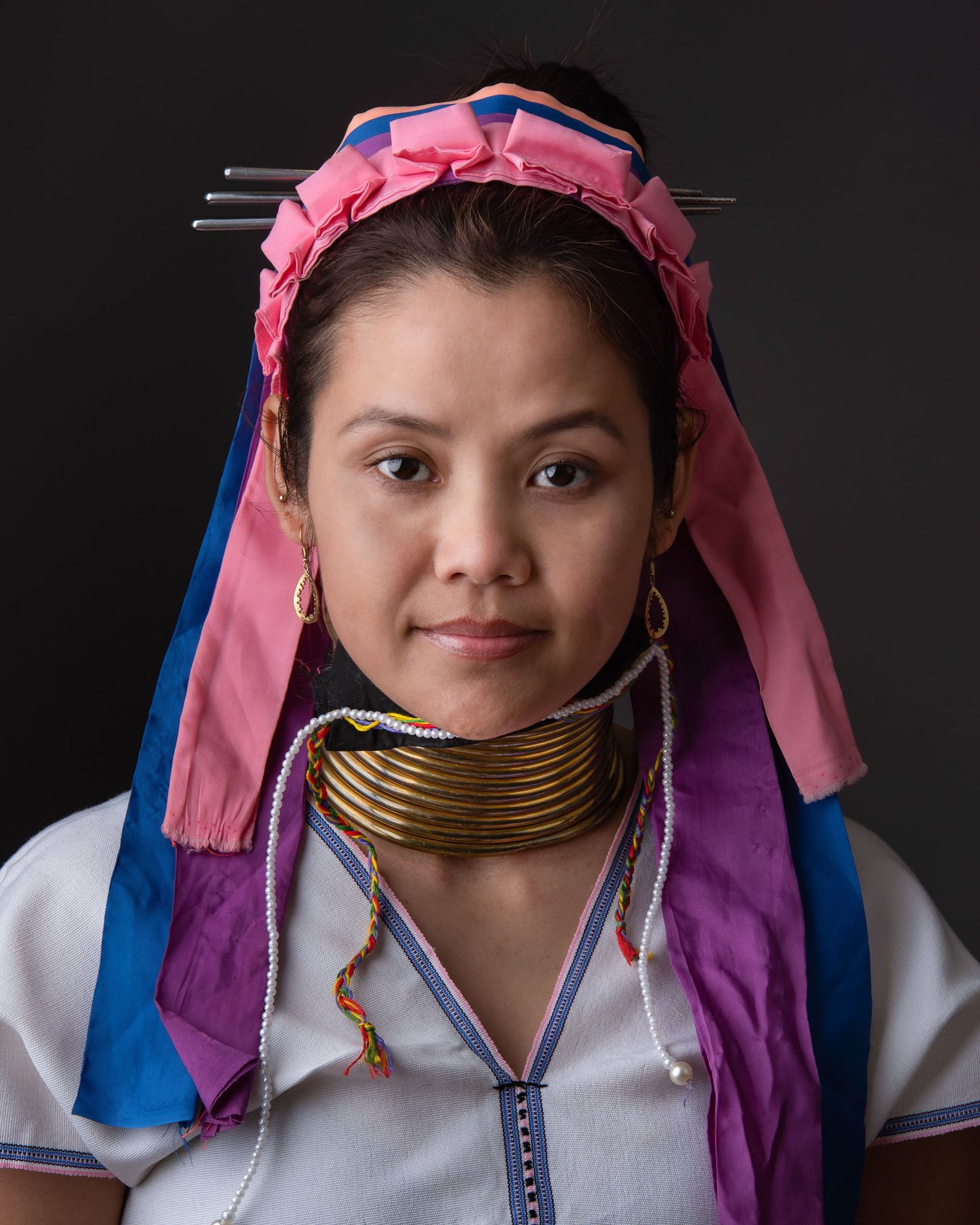 Traditional long-necked tribal woman wearing a pink headpiece and blue and purple garments against dark background.