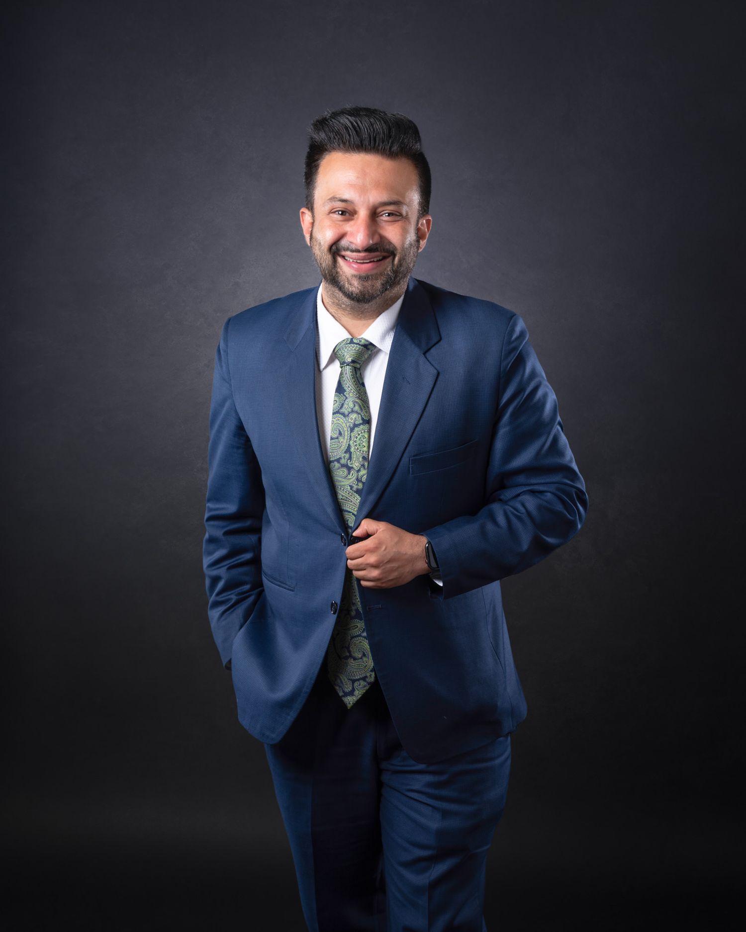 Professional business portrait of someone in a navy suit and floral tie smiling warmly against a dark background.