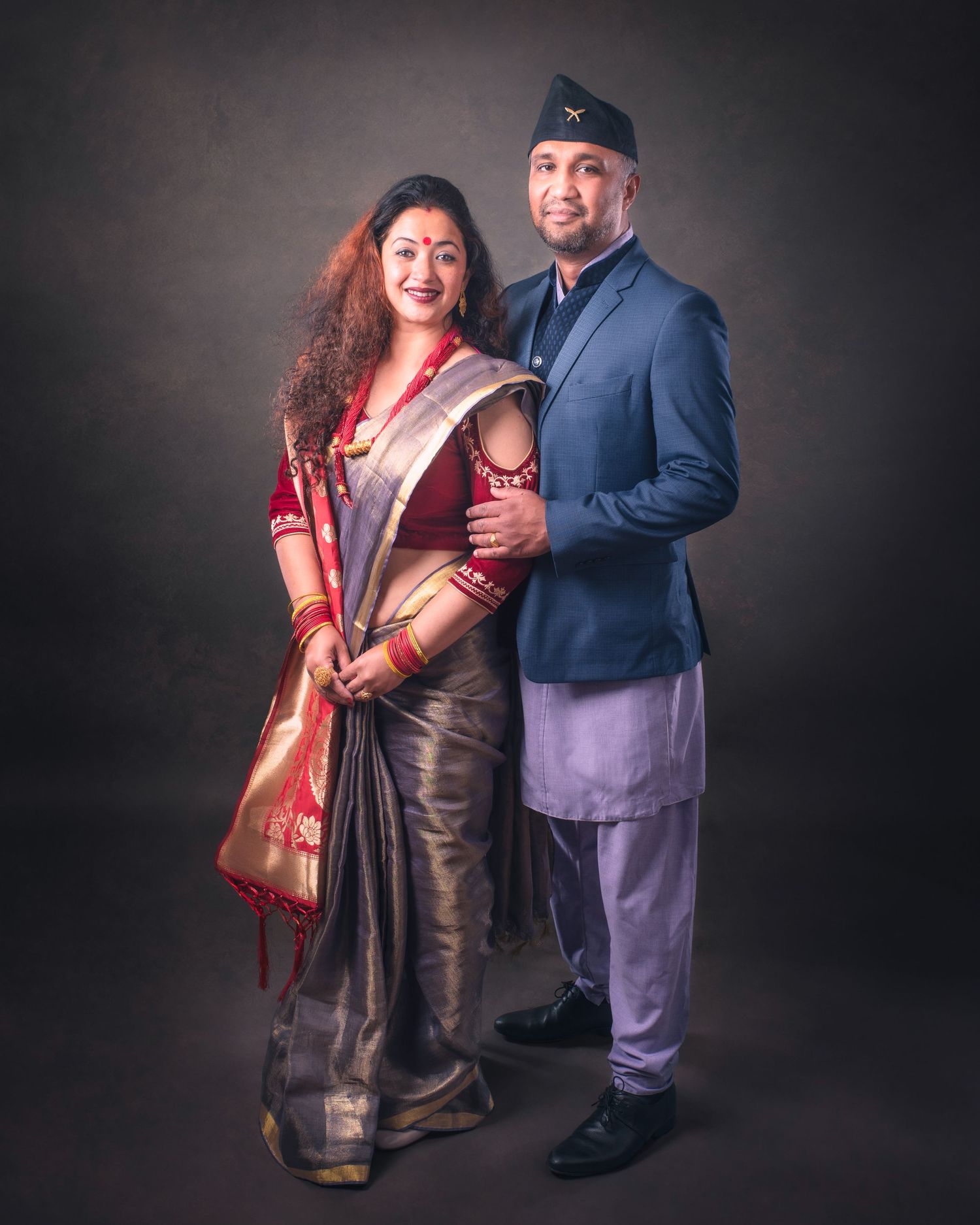 A couple poses together in traditional South Asian attire against a dark backdrop in a formal portrait setting.