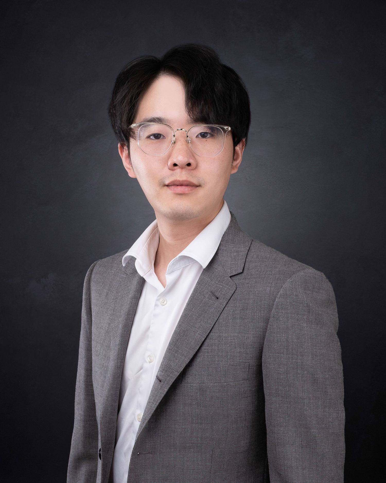 Professional headshot of person in grey suit jacket and white dress shirt against dark grey backdrop.