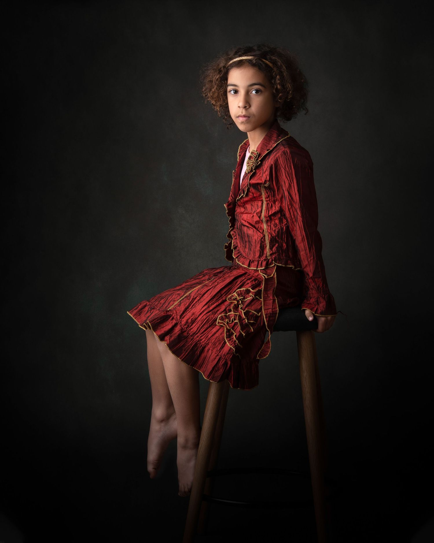 Artistic dark portrait of a young person in a flowing red dress sitting on a stool against a shadowy background.
