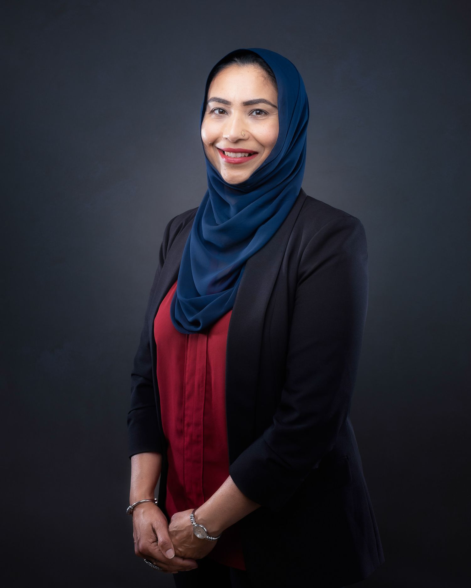 Professional portrait of someone in a navy hijab, red blouse and black blazer against a dark background.