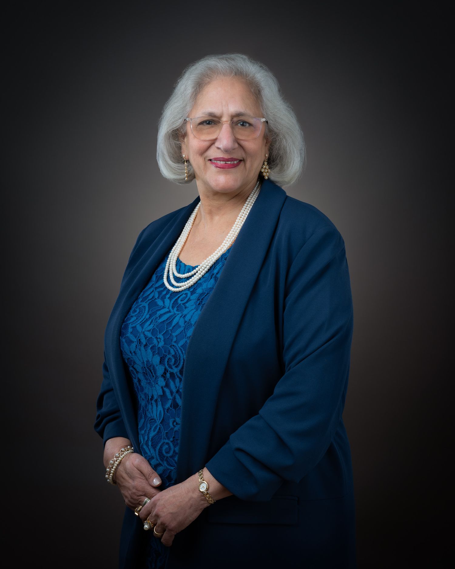 Professional portrait of a person in a navy blazer and blue patterned top with layered pearl necklaces against dark background.