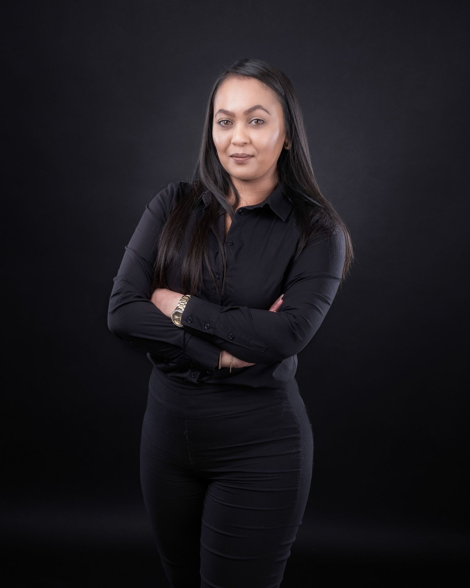 Professional portrait of someone standing against a dark background wearing all black attire with arms crossed.
