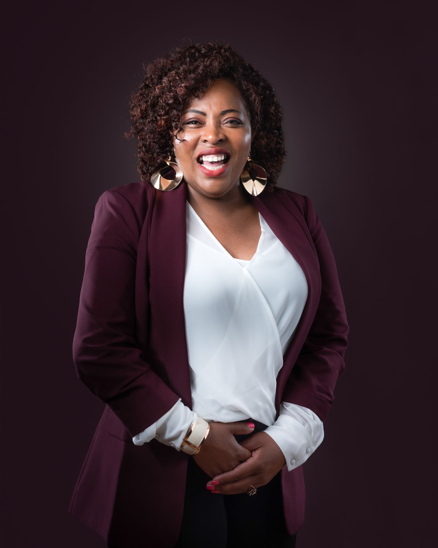 Professional studio portrait with someone wearing a burgundy blazer, white blouse and gold earrings against dark background.