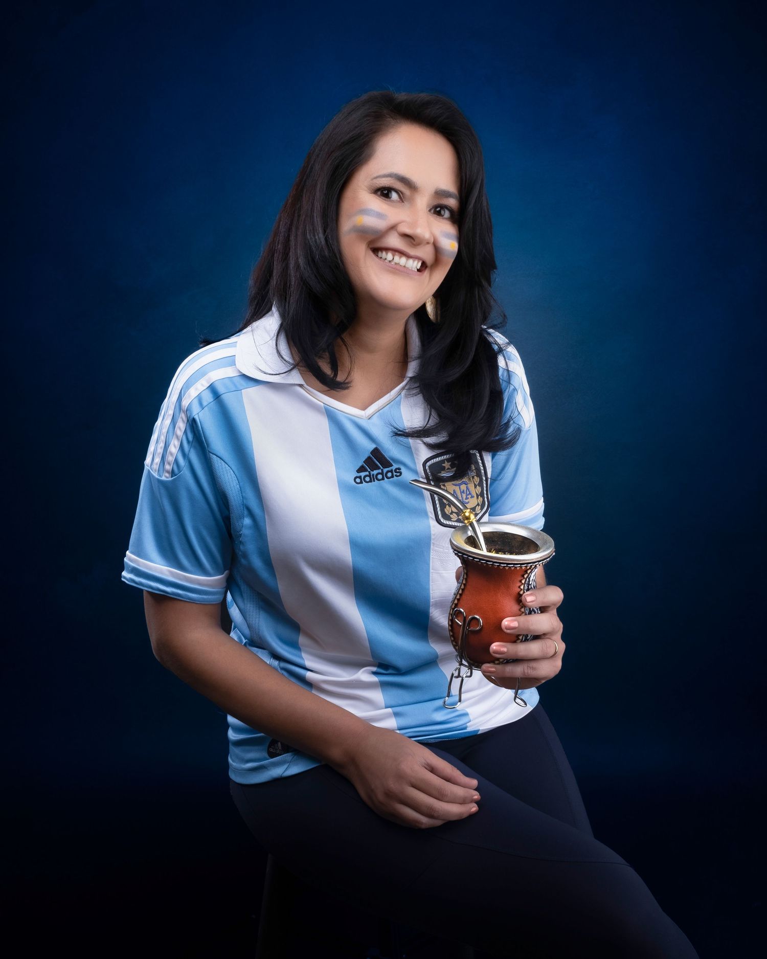 Soccer fan wearing Argentina's blue and white striped jersey smiles while holding mate drink against dark background.