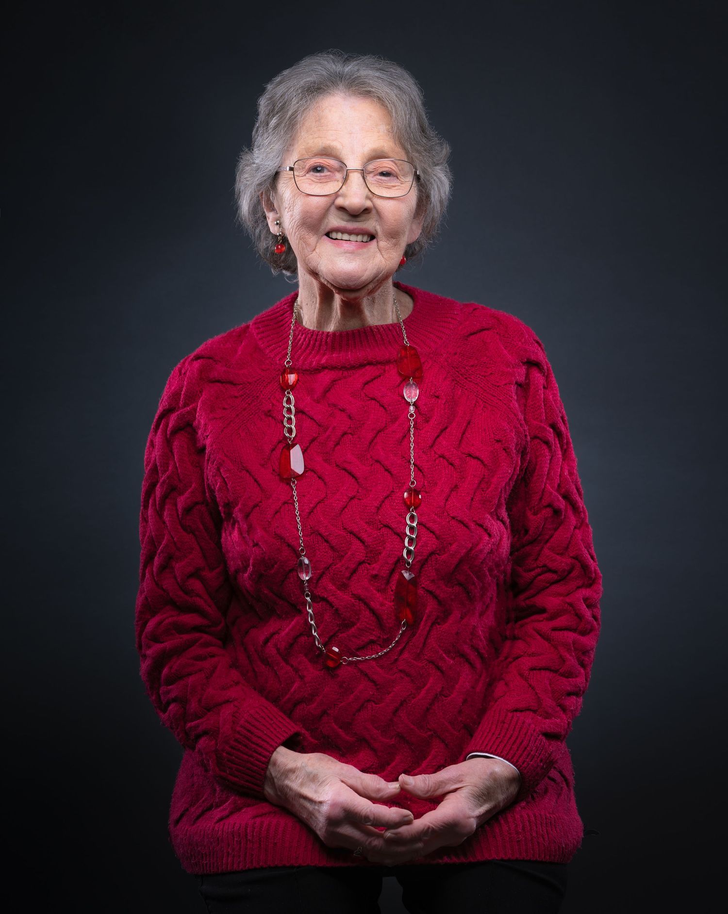 An elderly person wearing a red knit sweater and long beaded necklace poses against a dark background in a studio portrait.