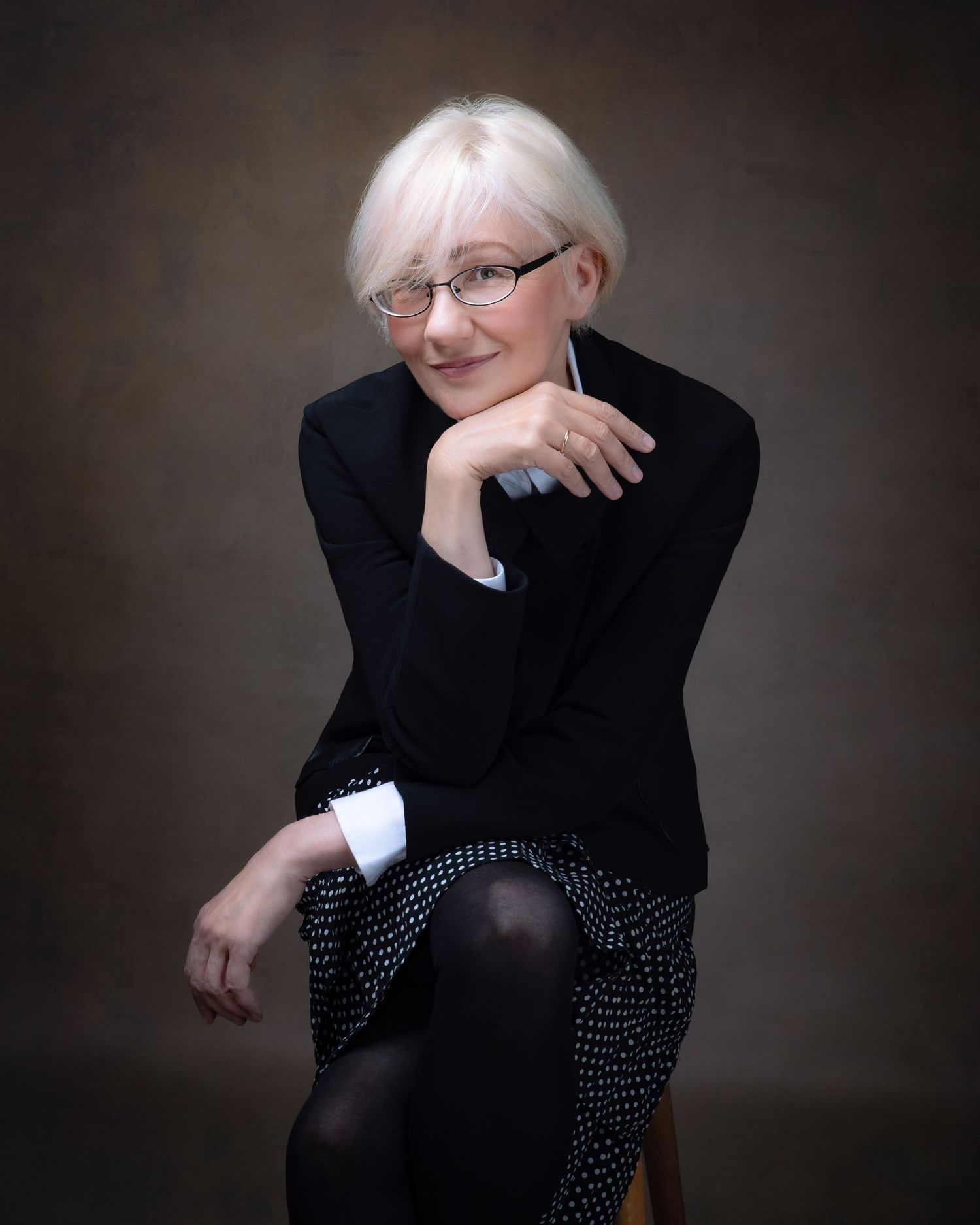 A sophisticated portrait featuring someone in a black blazer and polka dot skirt seated against a dark backdrop.