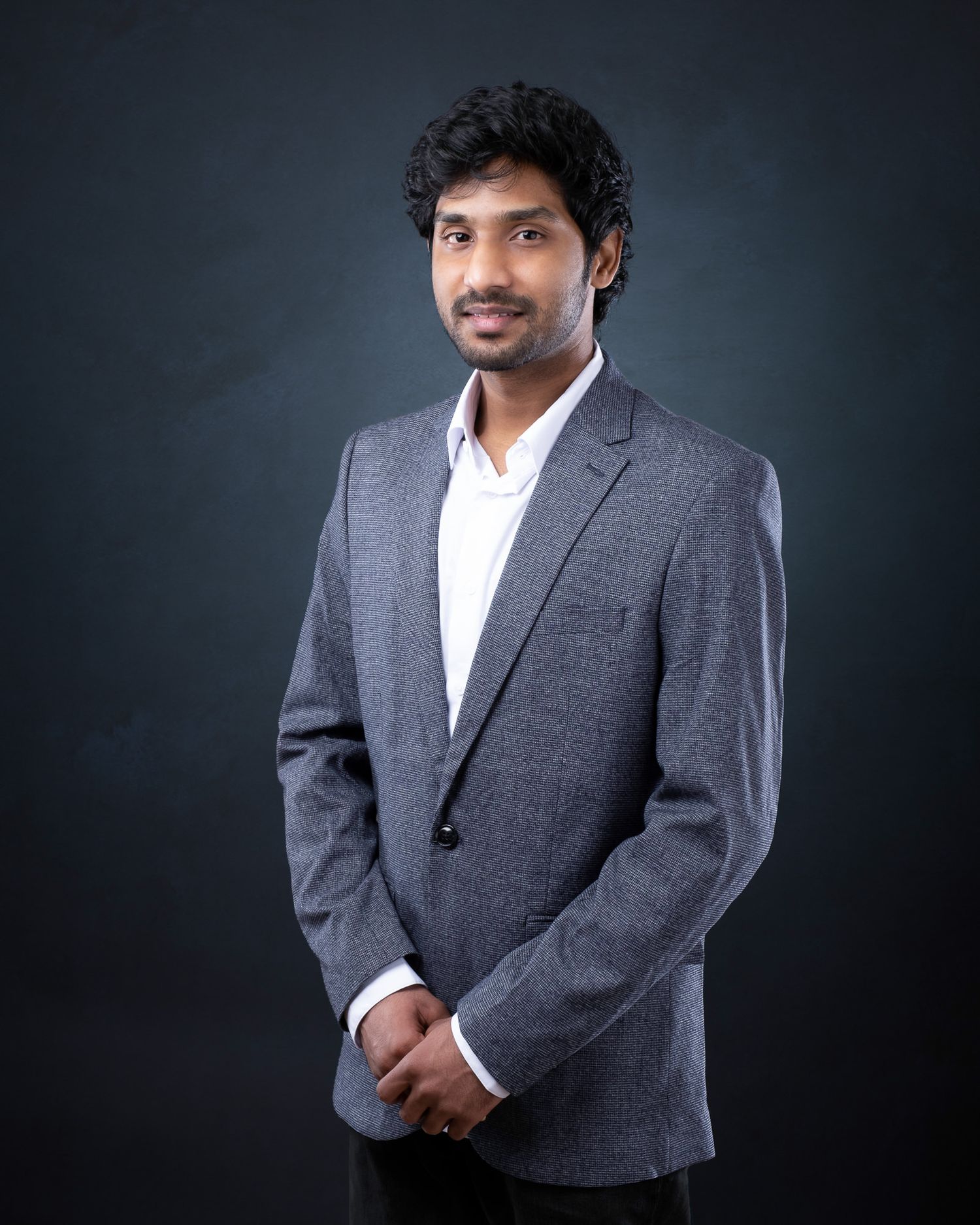Professional headshot of a young professional in a grey suit jacket and white shirt against dark background.