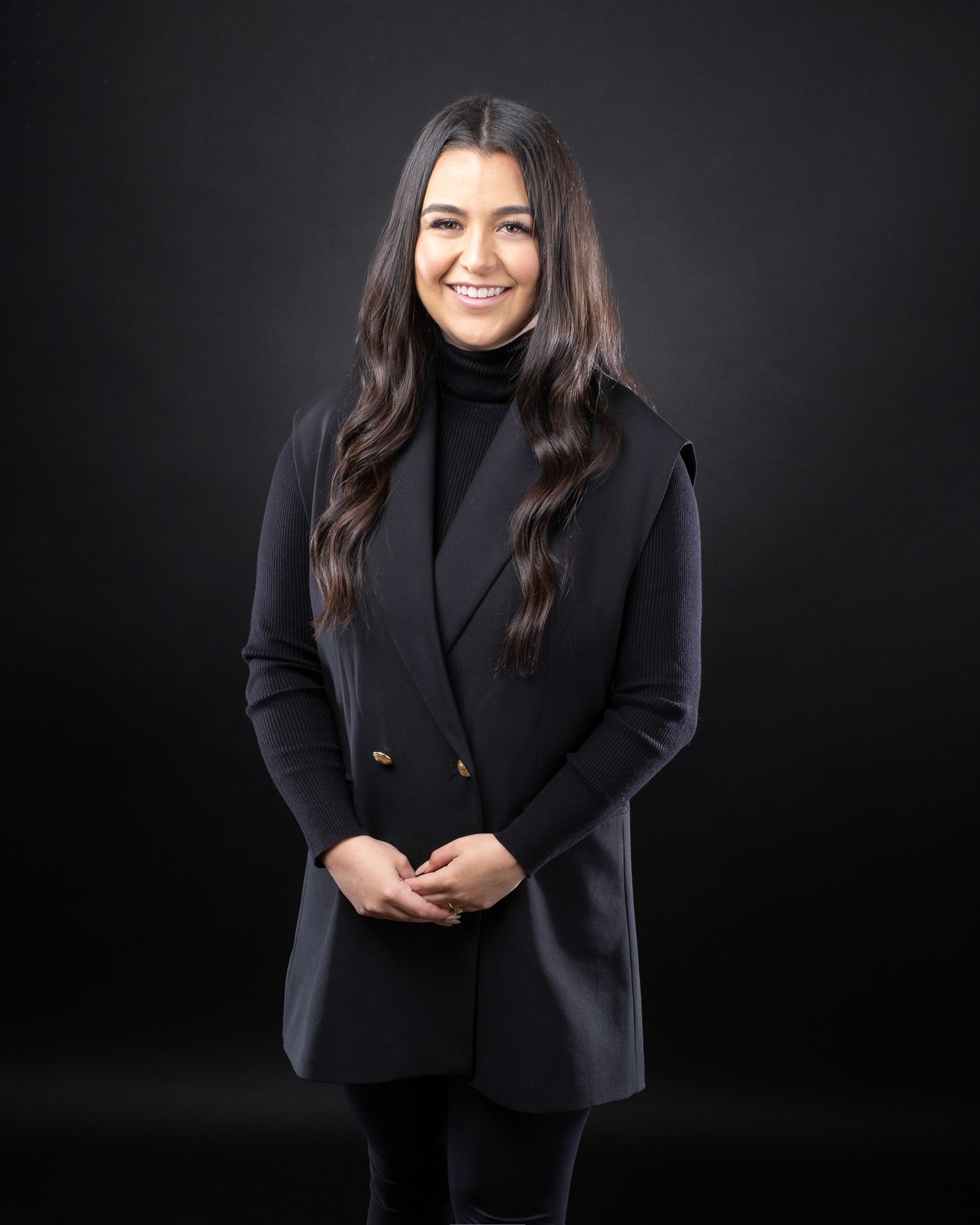Professional studio portrait on black background showing person in dark turtleneck and blazer with long dark hair.