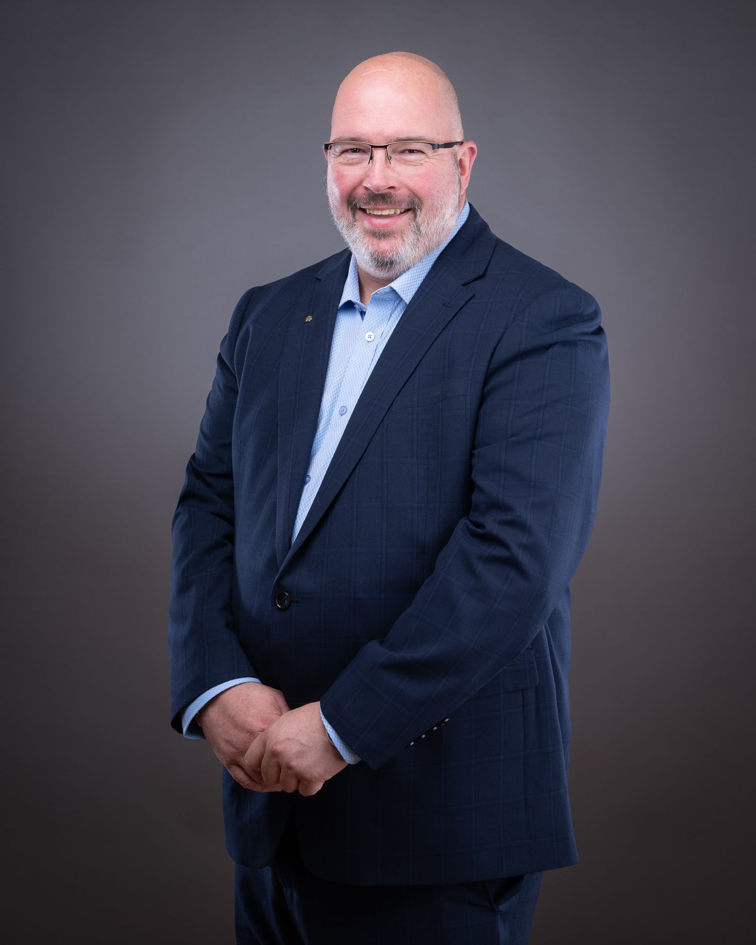 Professional business portrait of a person in a navy suit and light blue shirt smiling warmly against a dark background.