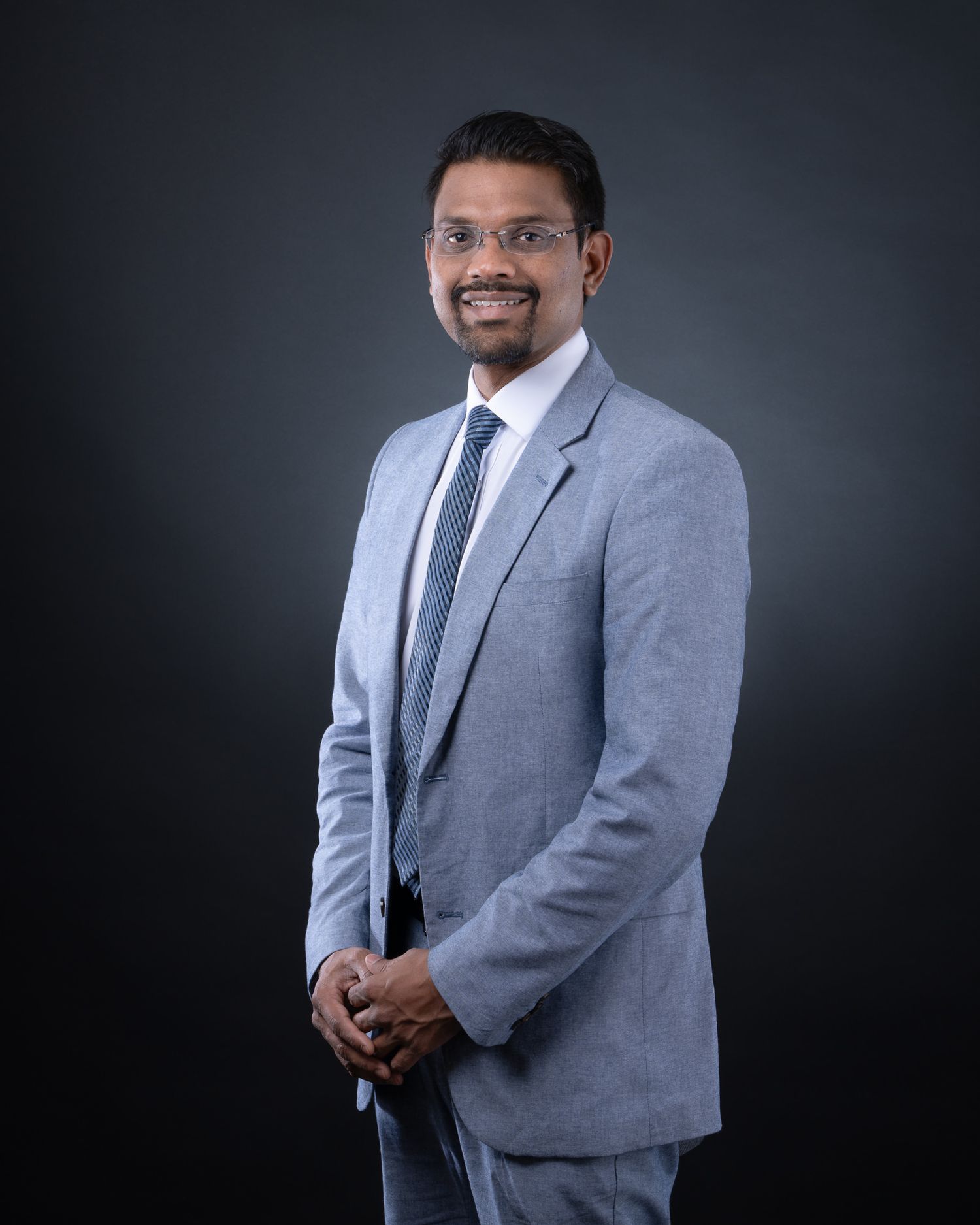 Professional portrait of a person in a grey suit and blue tie smiling against a dark background.