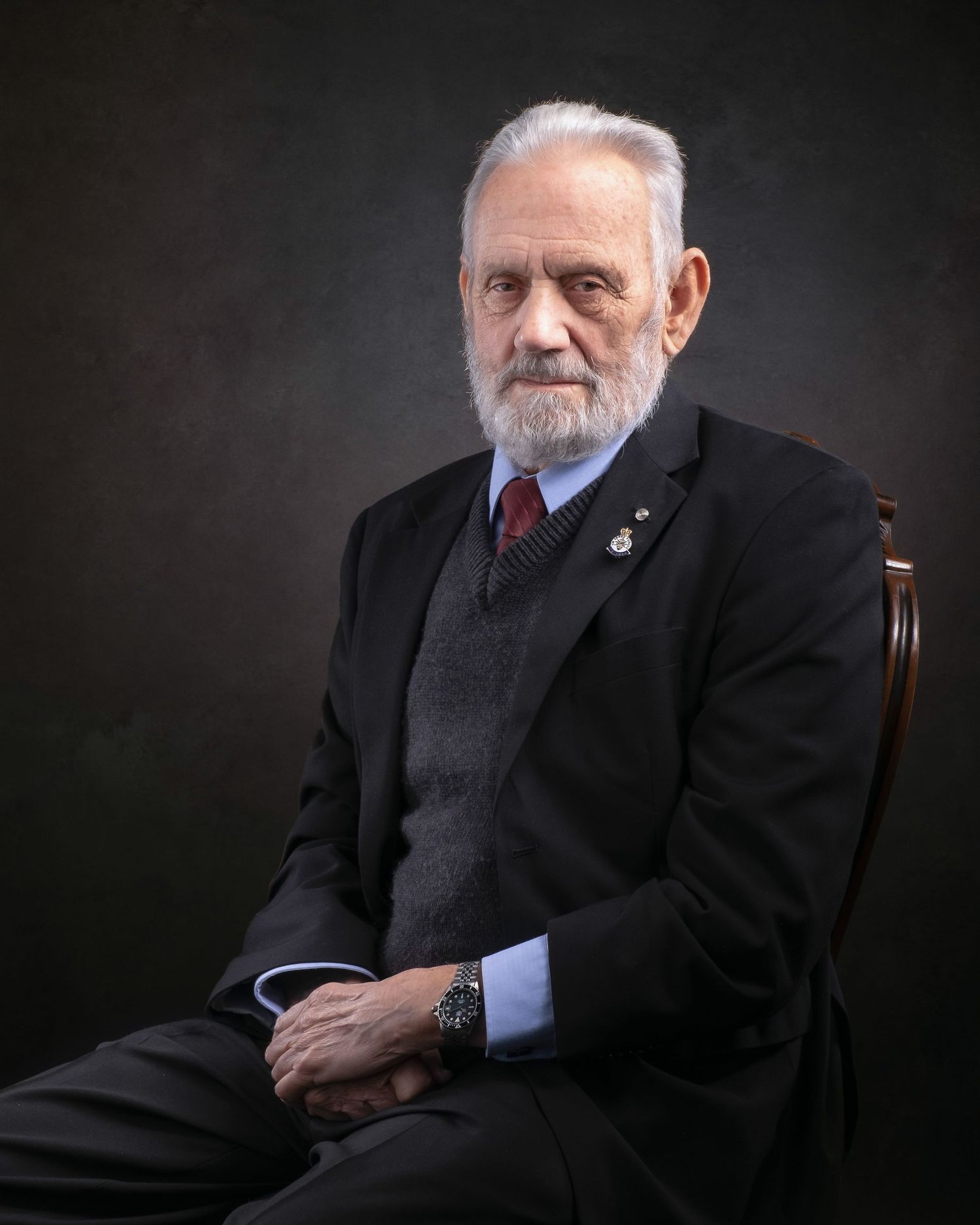 Professional studio portrait against dark background of an elderly individual in formal attire with a beard and gray hair.