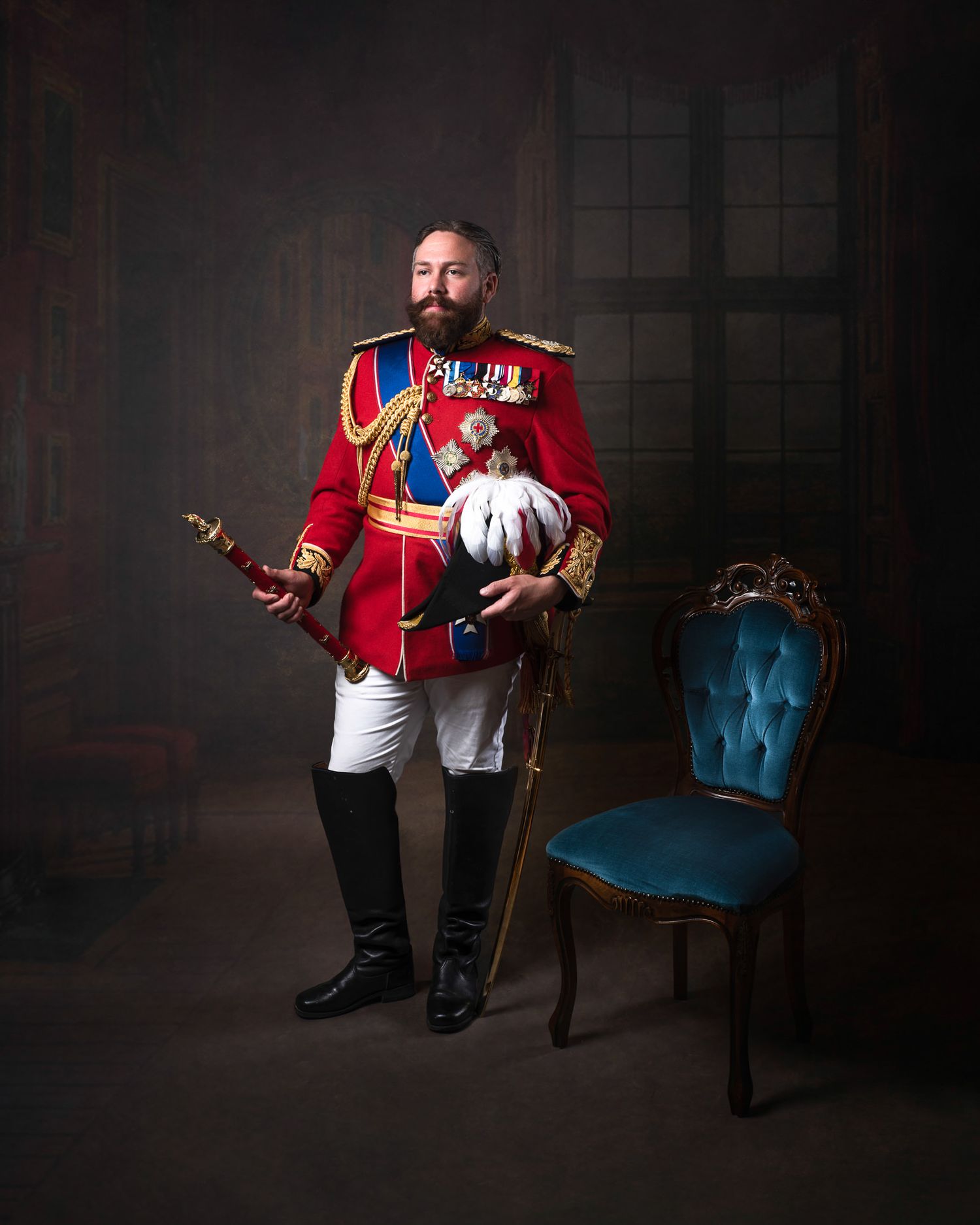 A military officer in ceremonial red dress uniform with medals stands beside a teal upholstered chair in a formal portrait setting.
