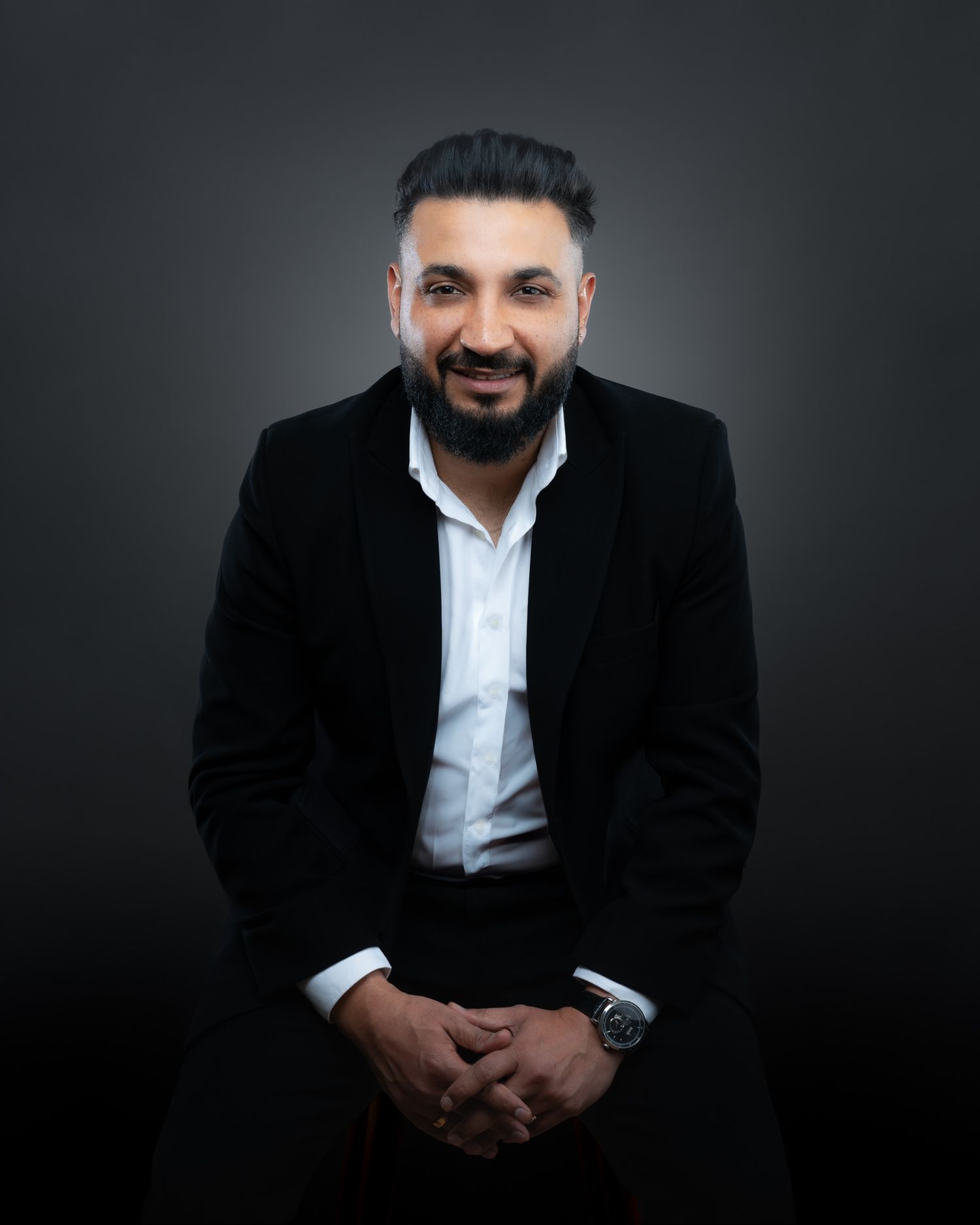 Professional studio portrait of a person in a black suit and white shirt seated against a dark background.
