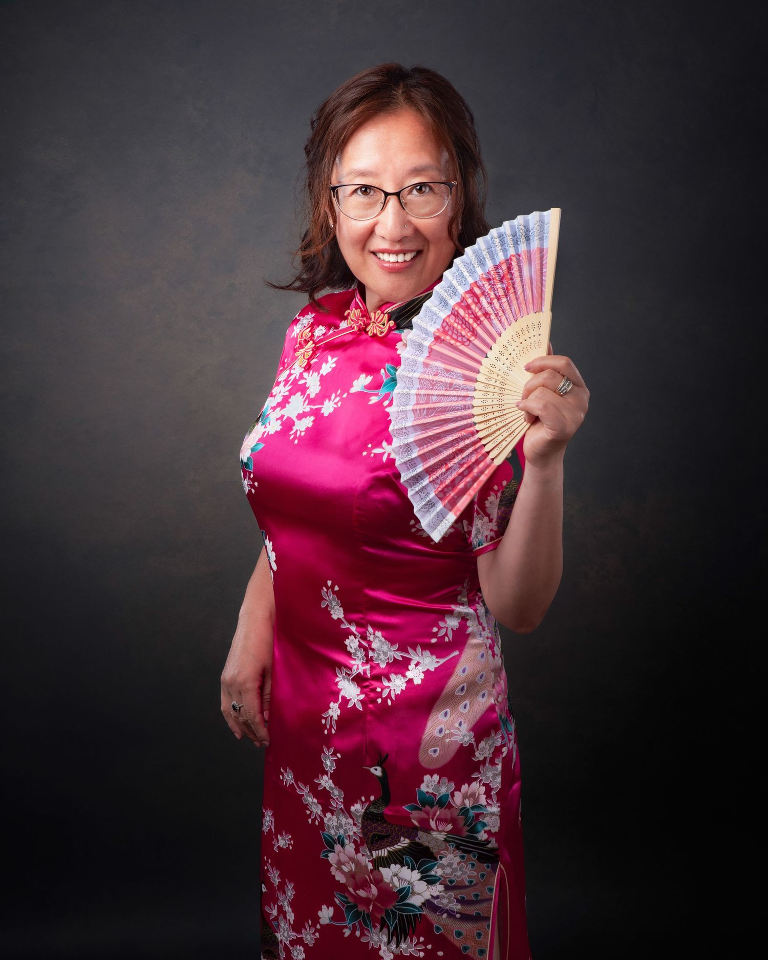 A person poses in a vibrant pink Asian-inspired dress with cherry blossoms while holding a decorative hand fan against dark backdrop.
