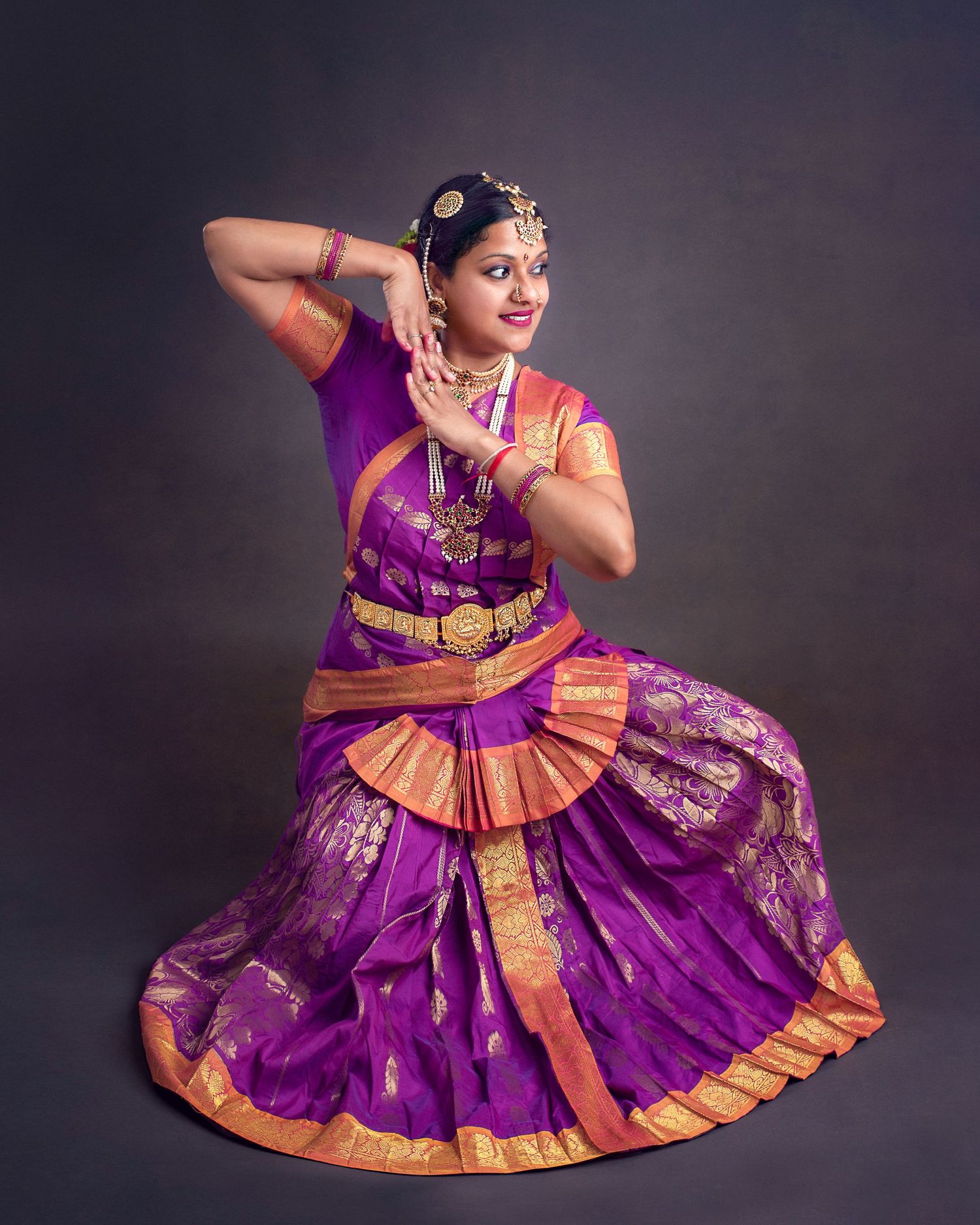 Classical Indian dancer in vibrant purple and gold sari performs traditional Bharatanatyam pose against dark backdrop.